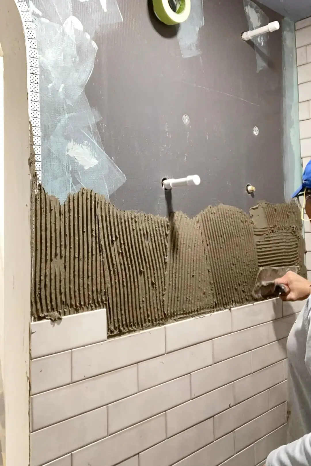 Close-up of a person using a trowel to spread adhesive onto a wall, preparing it for white subway tiles, with visible details of the tile pattern and the textured surface being worked on.