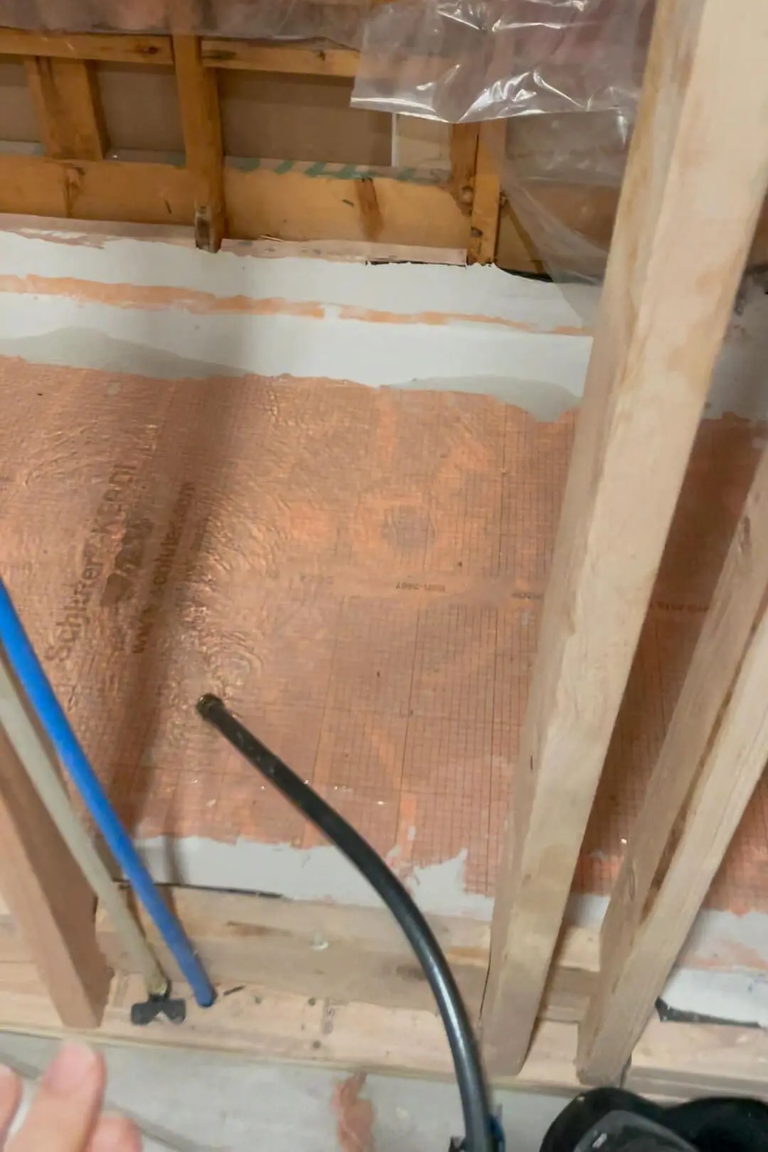 Close-up of a bathroom remodel with waterproofing membrane installed on the floor, visible through its distinctive orange color and grid pattern. Plumbing pipes extend up through the flooring, set against exposed wooden studs, indicating preparation for tile installation.