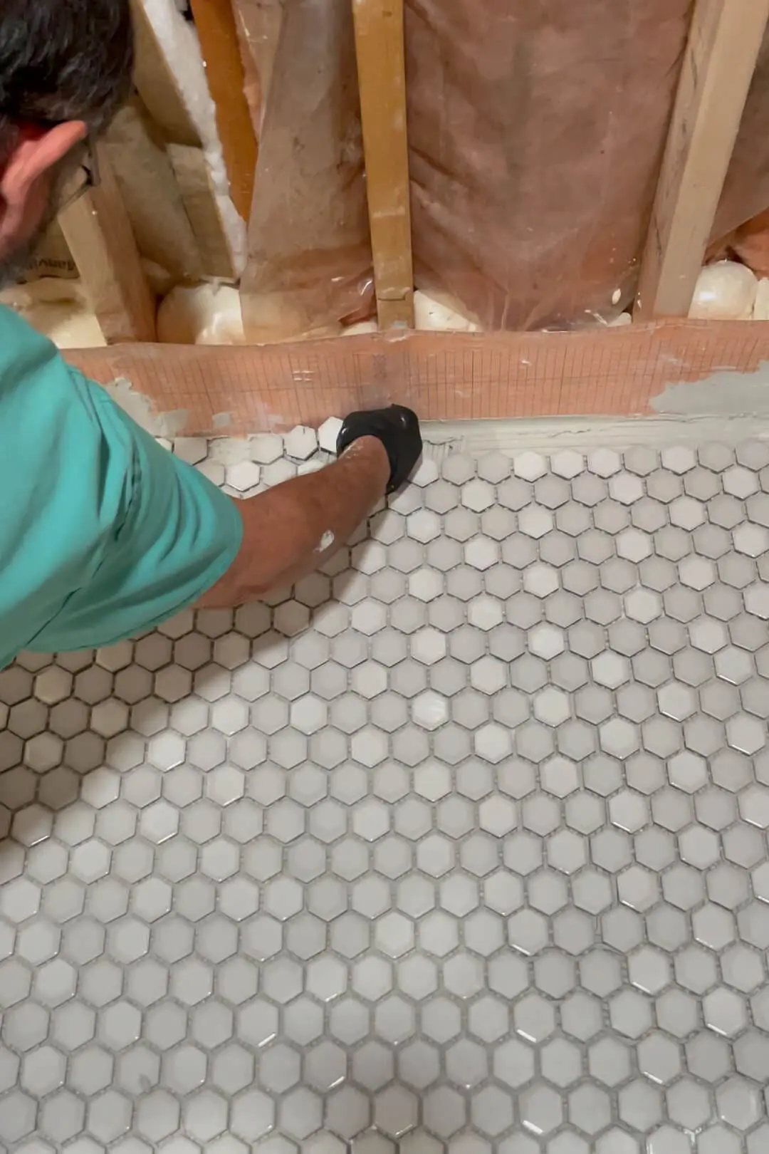 Close-up of a person installing small hexagon-shaped white tiles on a shower floor, with exposed wooden studs and insulation visible in the background, highlighting a DIY bathroom renovation project in progress.