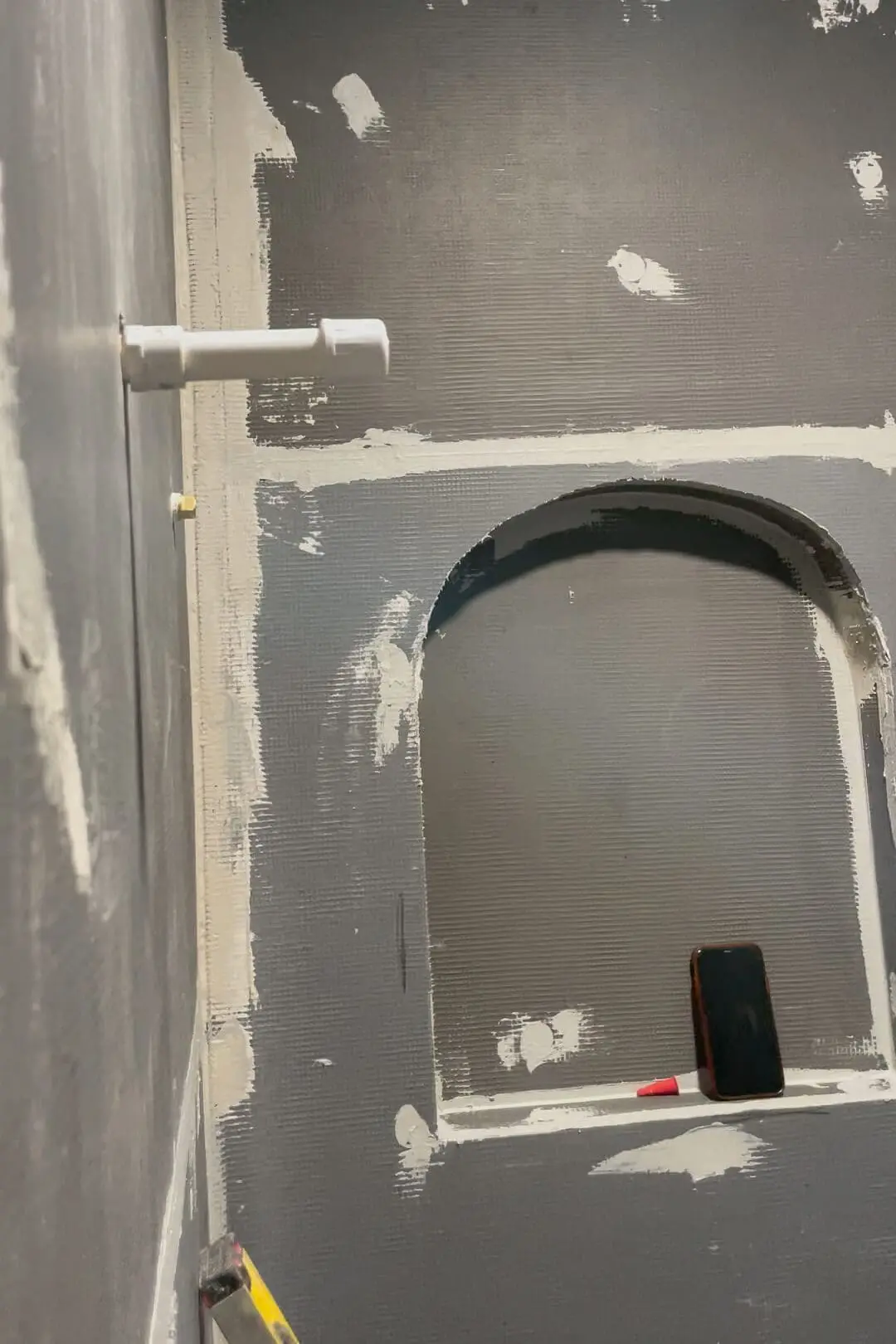 Close-up view of a prepared wall area with visible spackling patches, showing an installed white plumbing fixture and an outlet box, surrounded by a smooth, freshly applied primer coat, ready for the next steps in the bathroom renovation.