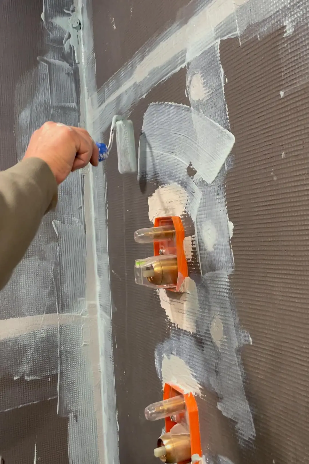 Person using a small roller brush to apply primer on a bathroom wall, covering spackling and outlet box areas, with a focus on ensuring an even coating on the wall surface to prepare it for tiling.
