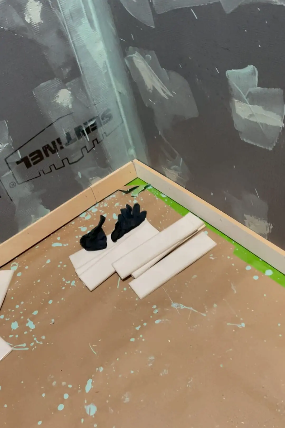 Close-up view of a tiled shower preparation area with black gloves and white subway tiles placed on a protective paper-covered floor. A ledger board has been installed to keep the tile level. The walls are primed with waterproofing material, and green painter's tape outlines the edges, ready for tile installation.