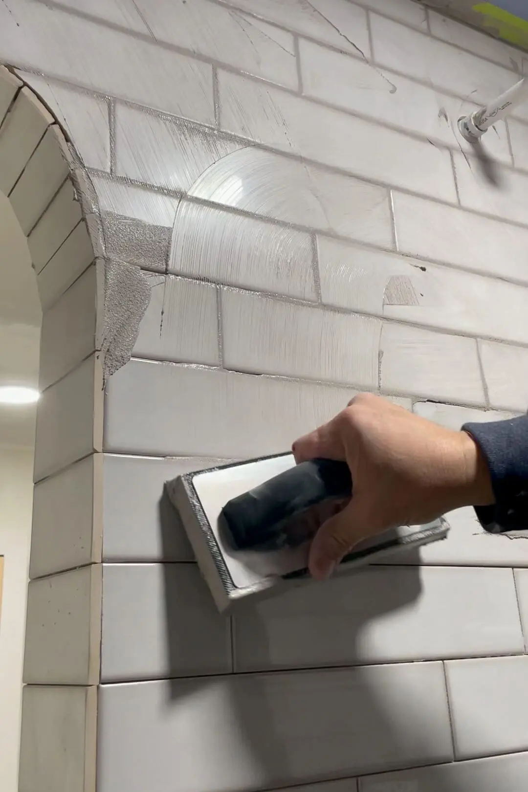 A close-up of a hand using a grout float to evenly apply grout to a subway-tiled shower, with focus on the curved arch detail being carefully finished.