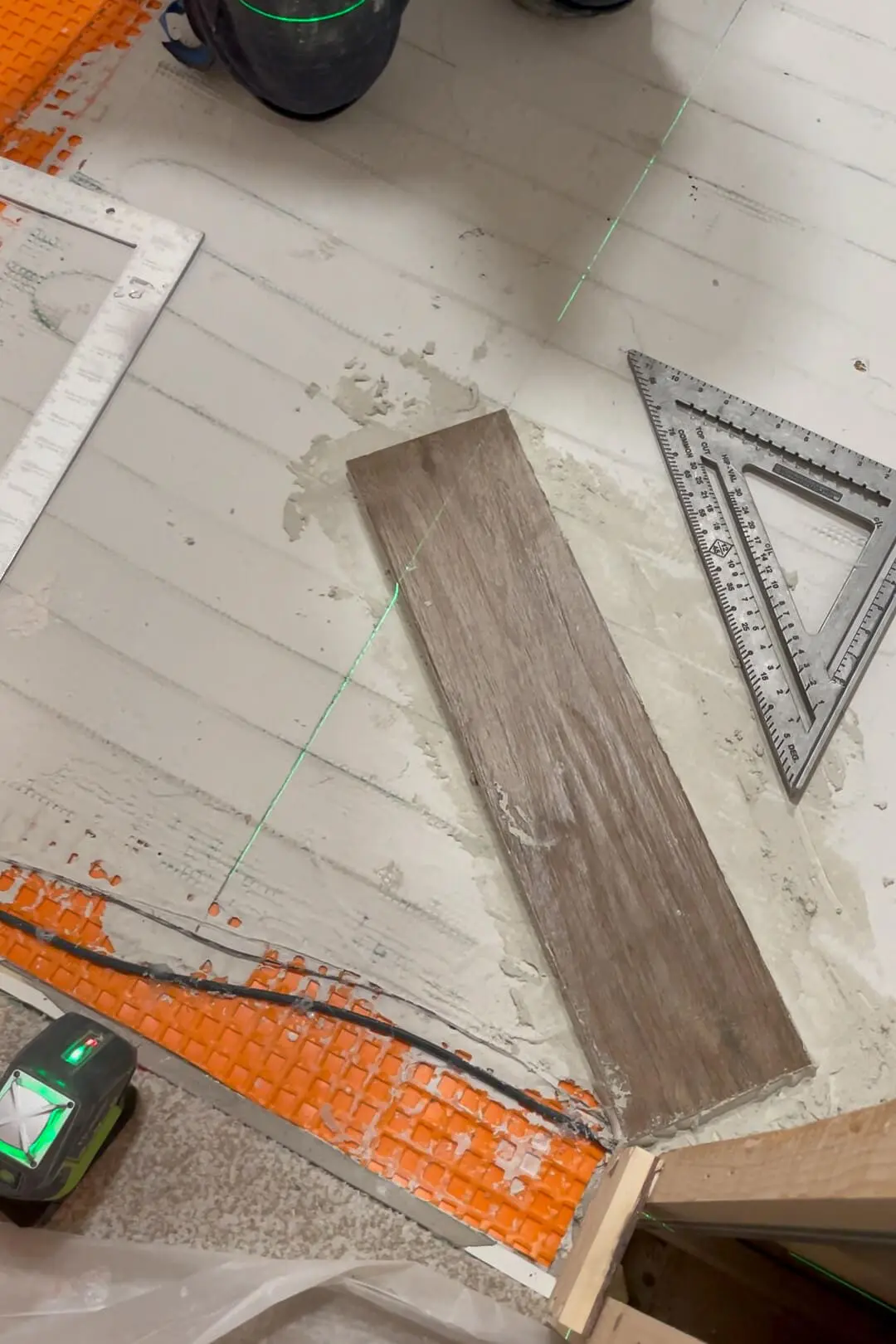 A piece of wood-look tile placed on a bathroom floor underlayment with visible laser guidelines, a t-square, and a Schluter-DITRA mat beneath, showcasing precise measurements for tile installation.