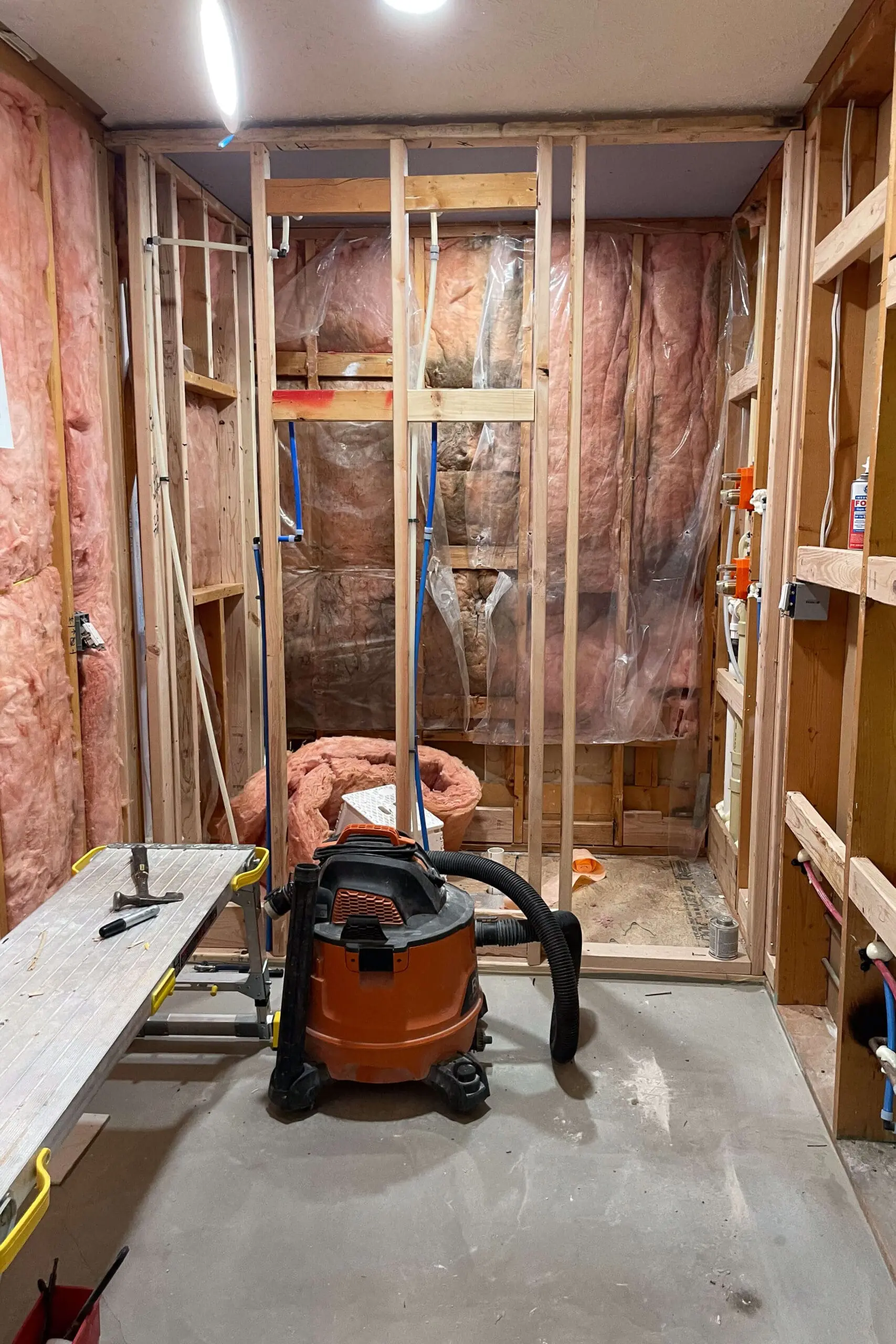 Bathroom under renovation with exposed framing and insulation, revealing the inner structure of the walls. The floor is concrete, and there is a shop vacuum and a workbench with tools, showcasing the early stages of a remodeling project.
