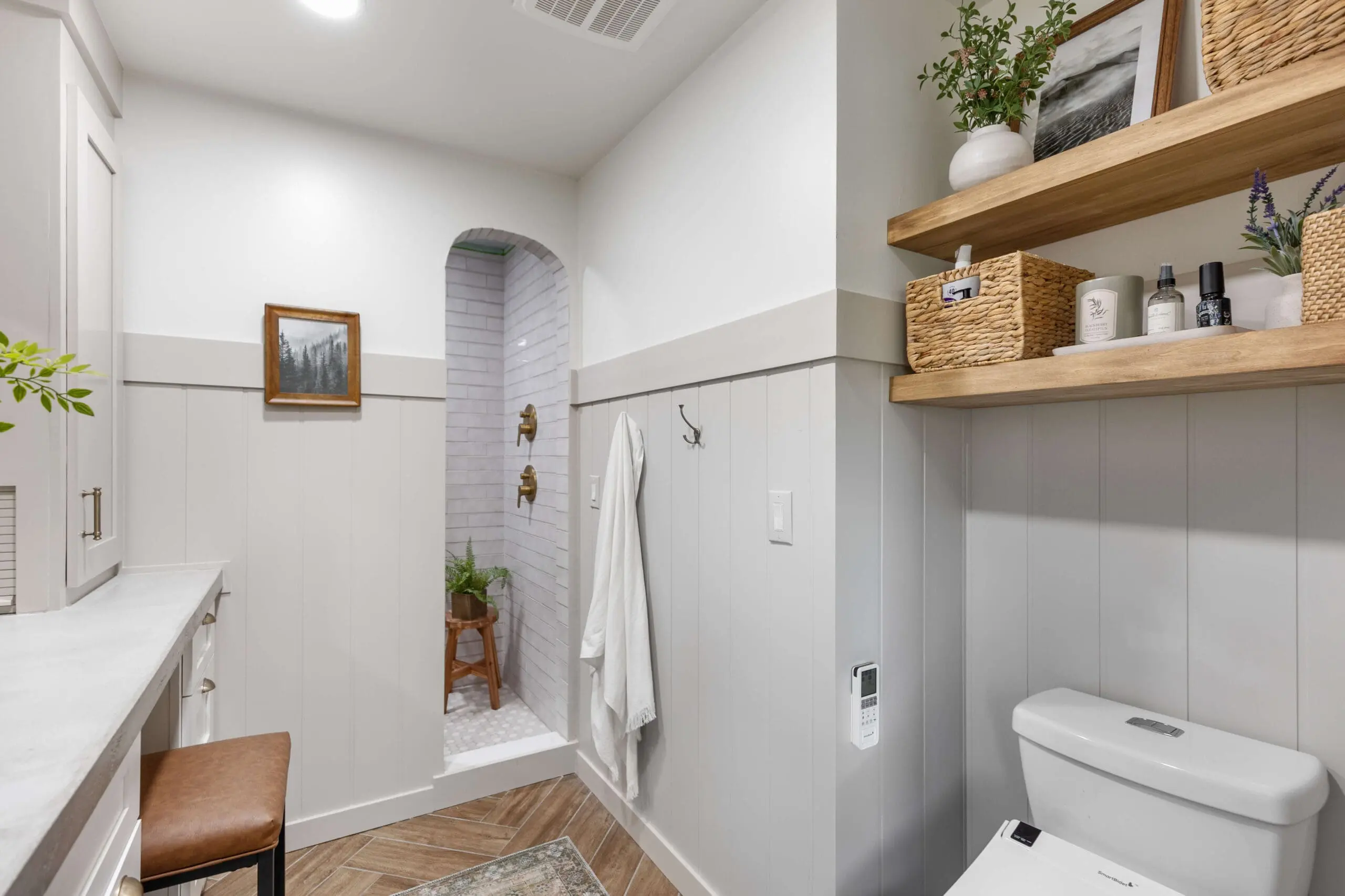 Bright and airy bathroom with an arched entryway leading into a tiled shower featuring matte gold fixtures and a small wooden stool topped with greenery. The main area includes warm wood floating shelves styled with baskets, plants, and decorative items above the toilet, while the walls are finished with half-height board and batten paneling. A soft neutral color palette and herringbone wood-look tile flooring create a cozy, spa-like atmosphere. A towel hangs from a hook, and a framed nature print adds a touch of character to the wall.
