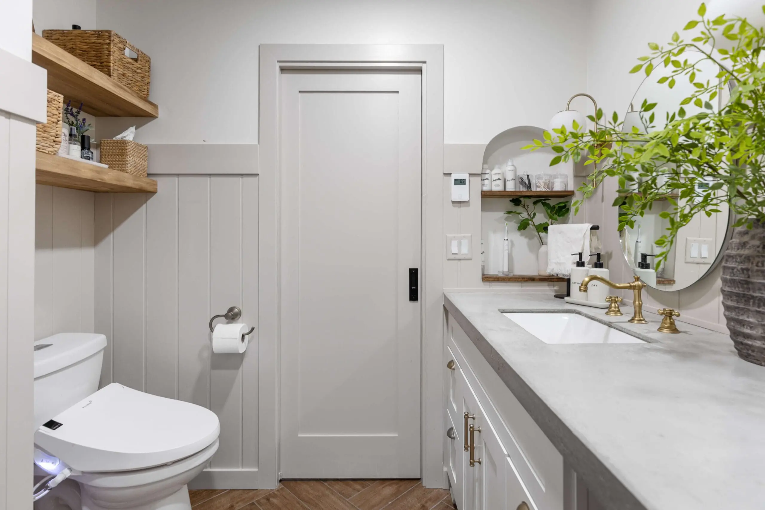 Modern farmhouse bathroom featuring a neutral palette with light gray walls and wainscoting. A sleek sliding barn door sits between built-in wood shelves with woven baskets and a concrete countertop with brass fixtures. A circular mirror is framed by greenery for a fresh, natural touch.