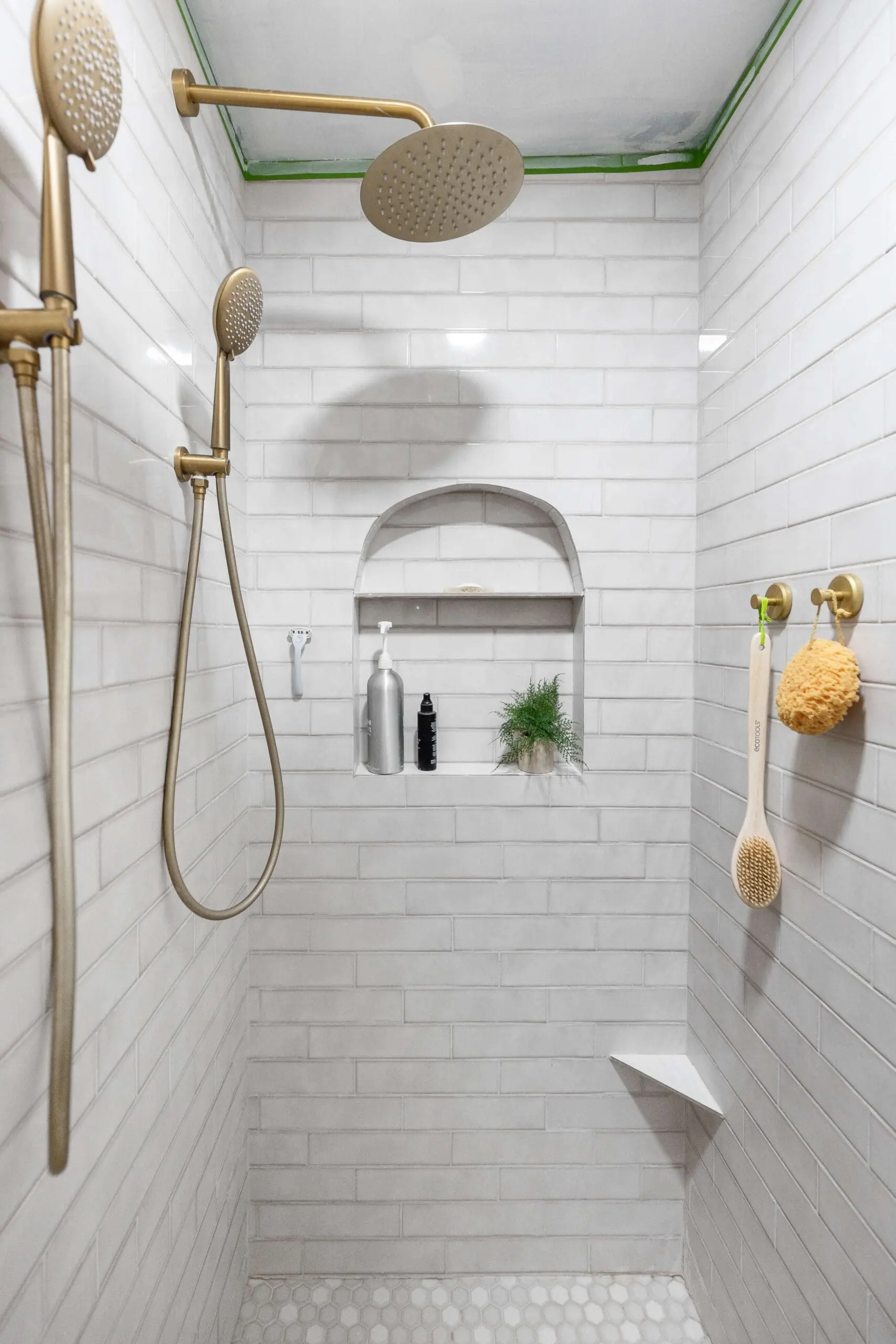 Luxurious subway tile shower with brass dual showerheads, including a rainfall and handheld shower fixture, offering a spa-like experience. The wall niche has an elegant arch and displays a small plant, silver soap dispenser, and black shampoo bottle for a minimalist look. The wall also features two brass hooks holding a natural sponge and wooden back scrubber, adding a warm, organic feel to the clean, white subway tile design.