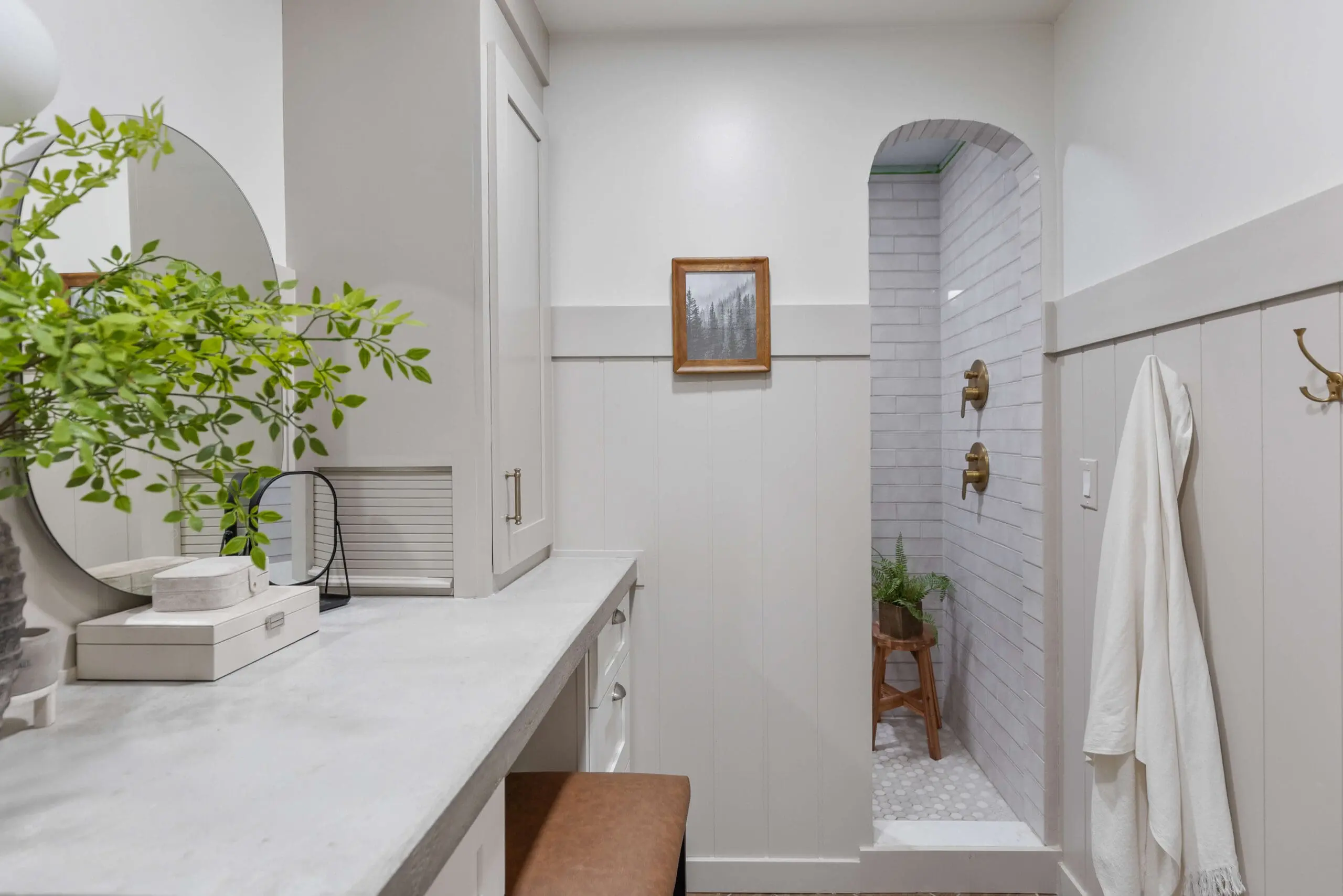 Modern bathroom with a soft, neutral palette and a subtle subway tile shower visible through an arched entryway. The shower has brass fixtures, and a wooden stool holding greenery enhances the spa vibe. The vanity area includes a round mirror, a green leafy plant, and stylish storage boxes, all sitting on a concrete countertop for a blend of elegance and functionality. A simple framed art piece on the wall and a white towel hanging from a brass hook add finishing touches to this serene, inviting bathroom space.
