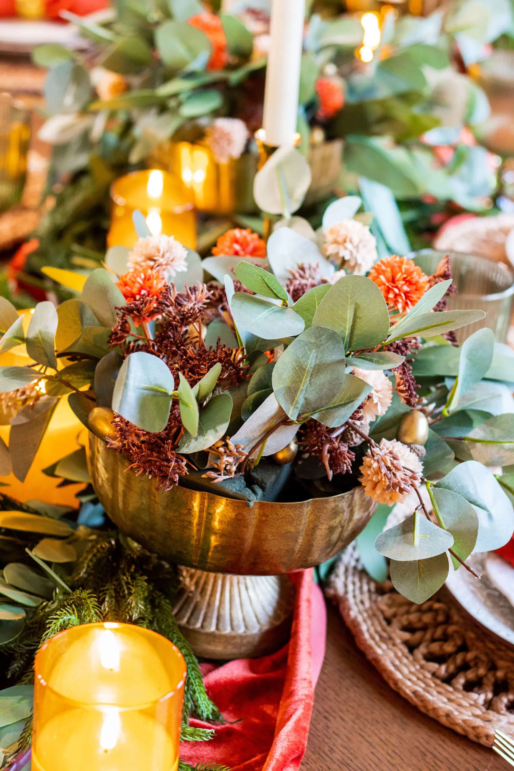 Thanksgiving table decor with brass candlesticks, green goblets, and greenery centerpiece, complemented by woven placemats and bright orange napkins for a seasonal touch.