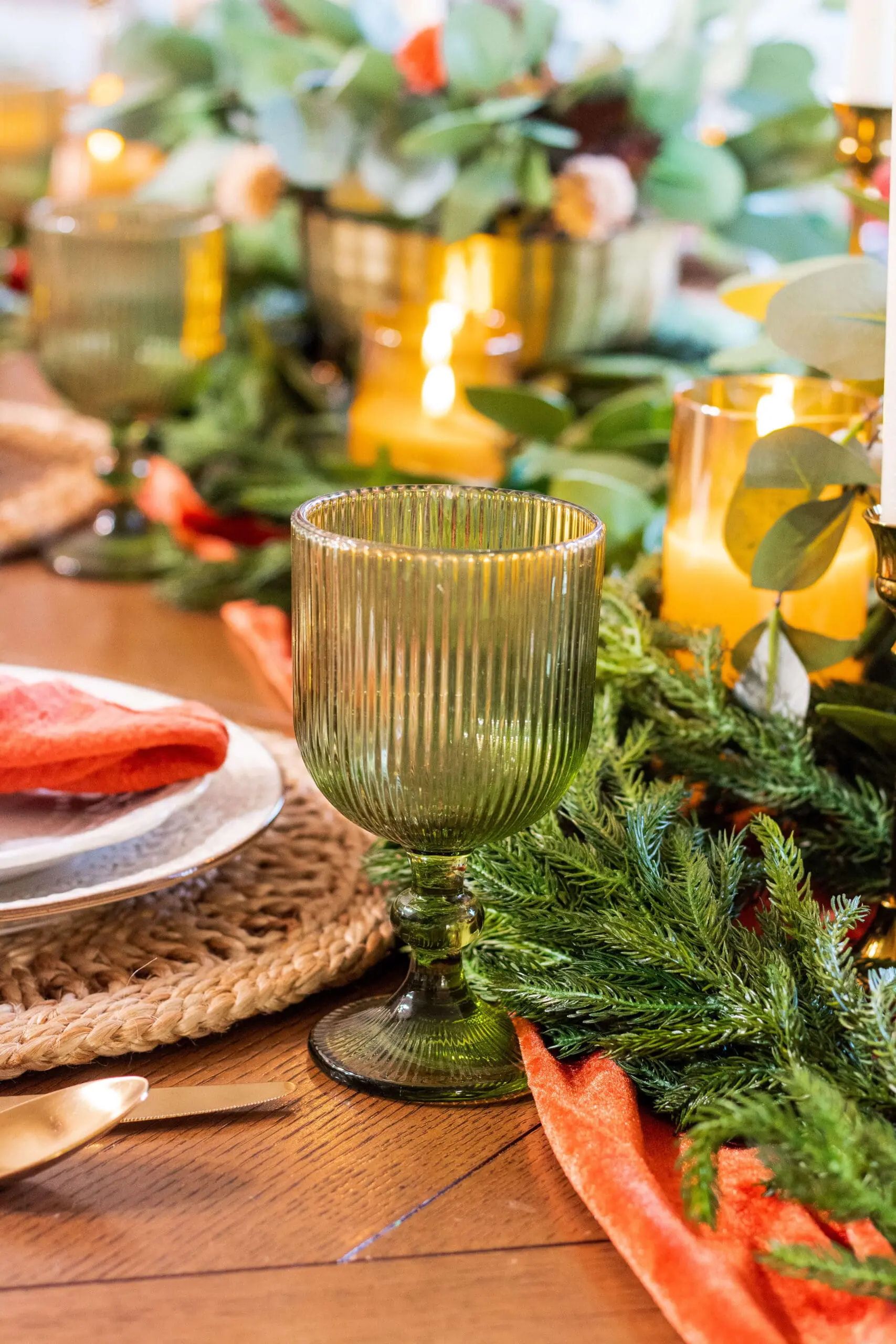 Green goblet glass beside a glowing candle on a Thanksgiving table decorated with rich greenery and an orange velvet runner, adding warmth to the holiday setting.
