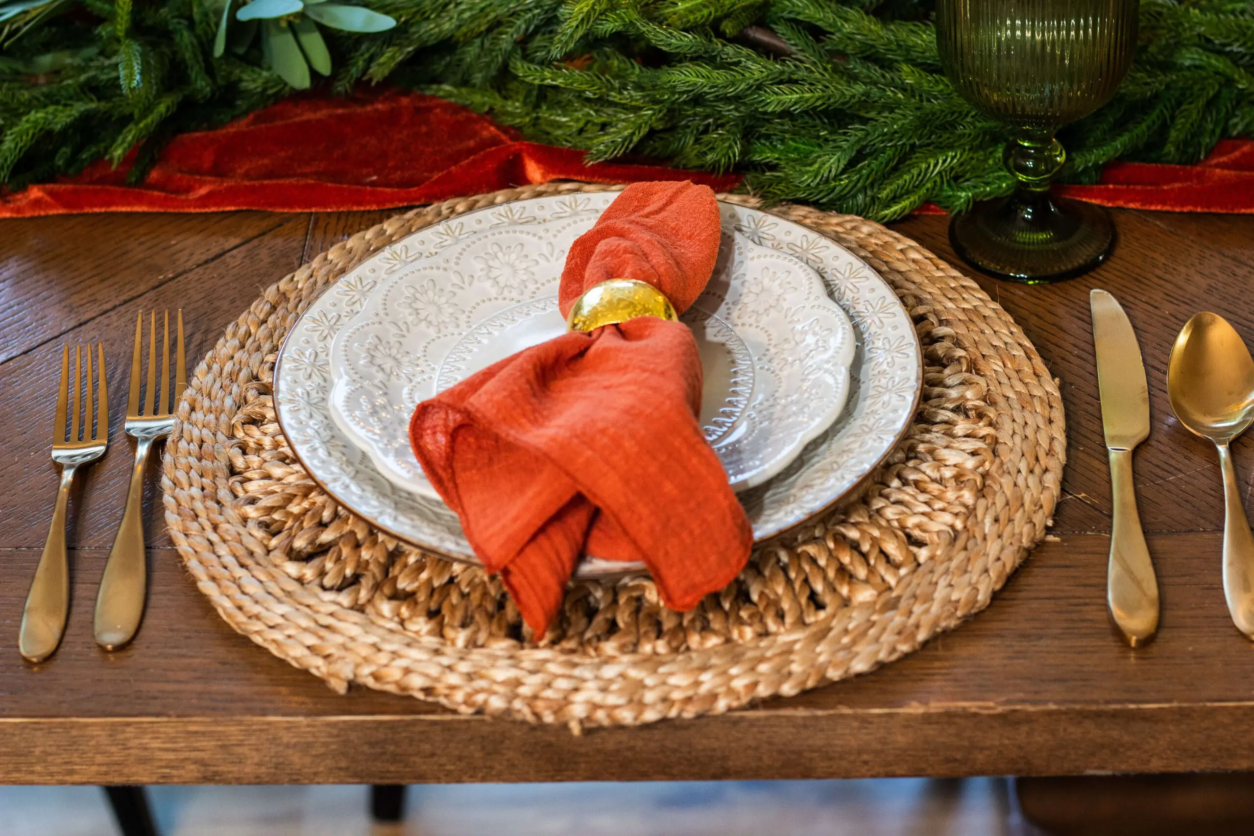 Place setting for Thanksgiving table decor with orange napkin wrapped in a gold napkin ring on a woven placemat, surrounded by greenery and warm autumnal accents.