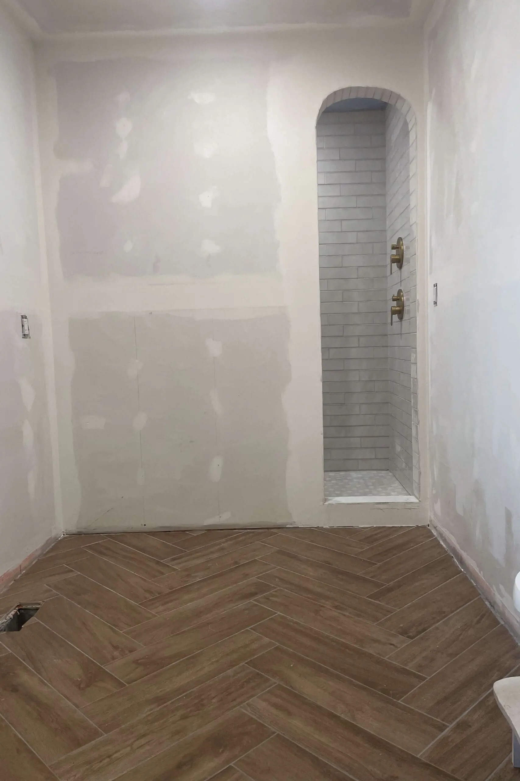 A nearly completed bathroom with freshly installed herringbone tile floor, smooth drywall ready for paint, and an elegant arched shower niche with white subway tiles and brass fixtures.