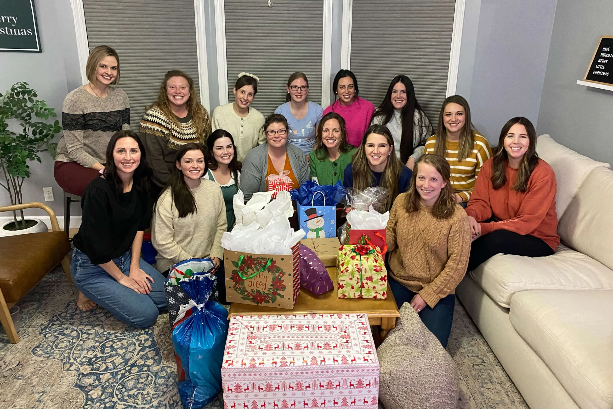 A group of fourteen women gathered together in a cozy living room for a holiday celebration. They’re smiling at the camera, surrounded by colorful, wrapped gifts placed on a table in front of them. The room is decorated with a festive sign on the wall that reads "Have Yourself a Merry Little Christmas." The women are dressed in warm, casual clothing, adding to the inviting and joyful holiday atmosphere.