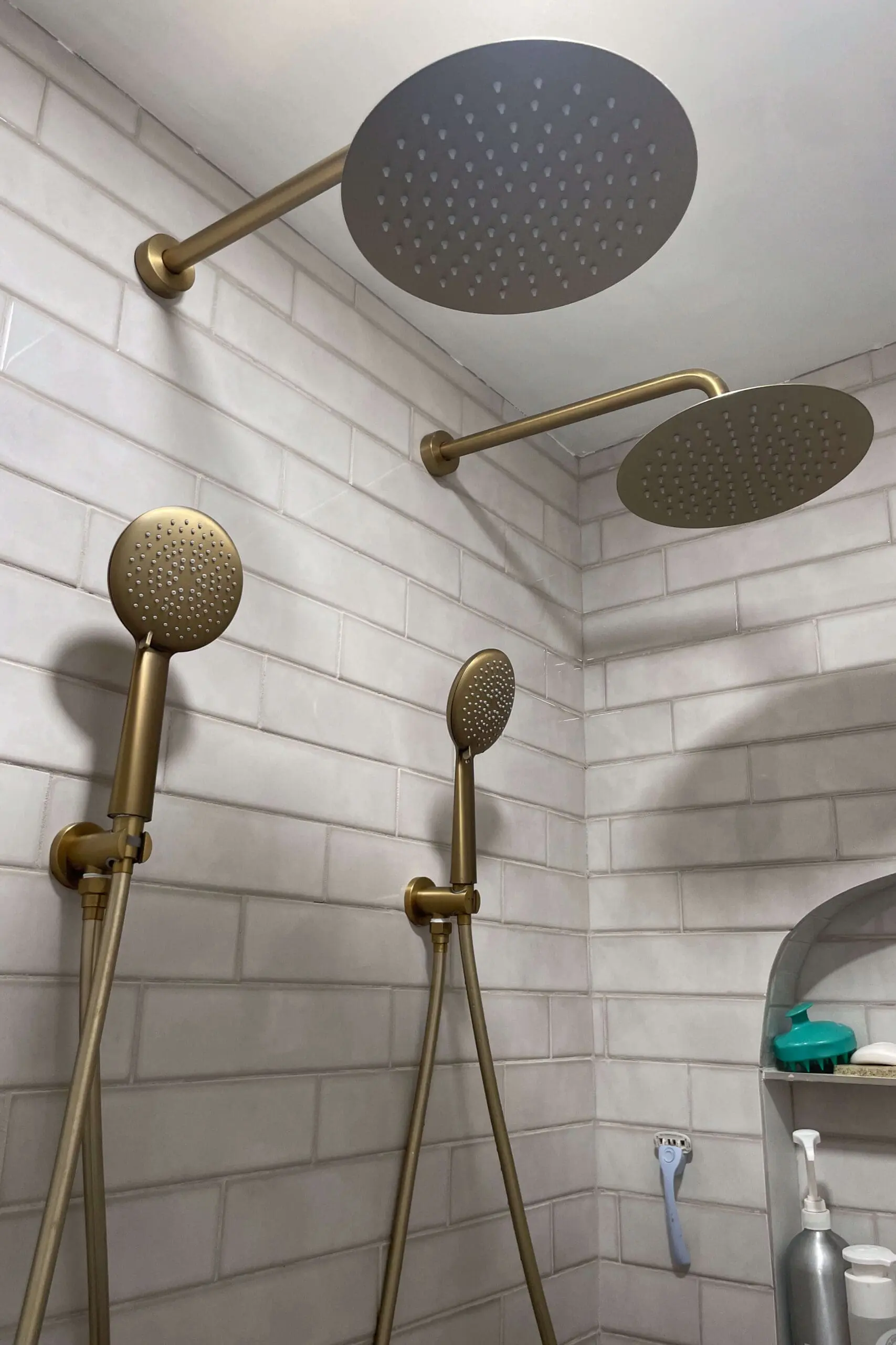 Dual rainfall shower heads and handheld shower wands in a matte gold finish, mounted on light gray subway tiles. The luxurious setup is enhanced by a shower niche with toiletries, exuding an upscale, hotel-inspired vibe.