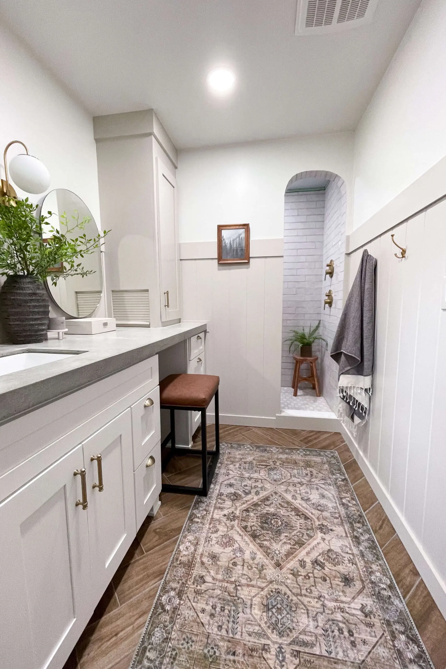 A DIY radiant floor heating bathroom project showcasing a bright, elegant space with a white vanity, gold accents, a patterned area rug, and wood-look tile flooring designed to distribute cozy warmth efficiently.