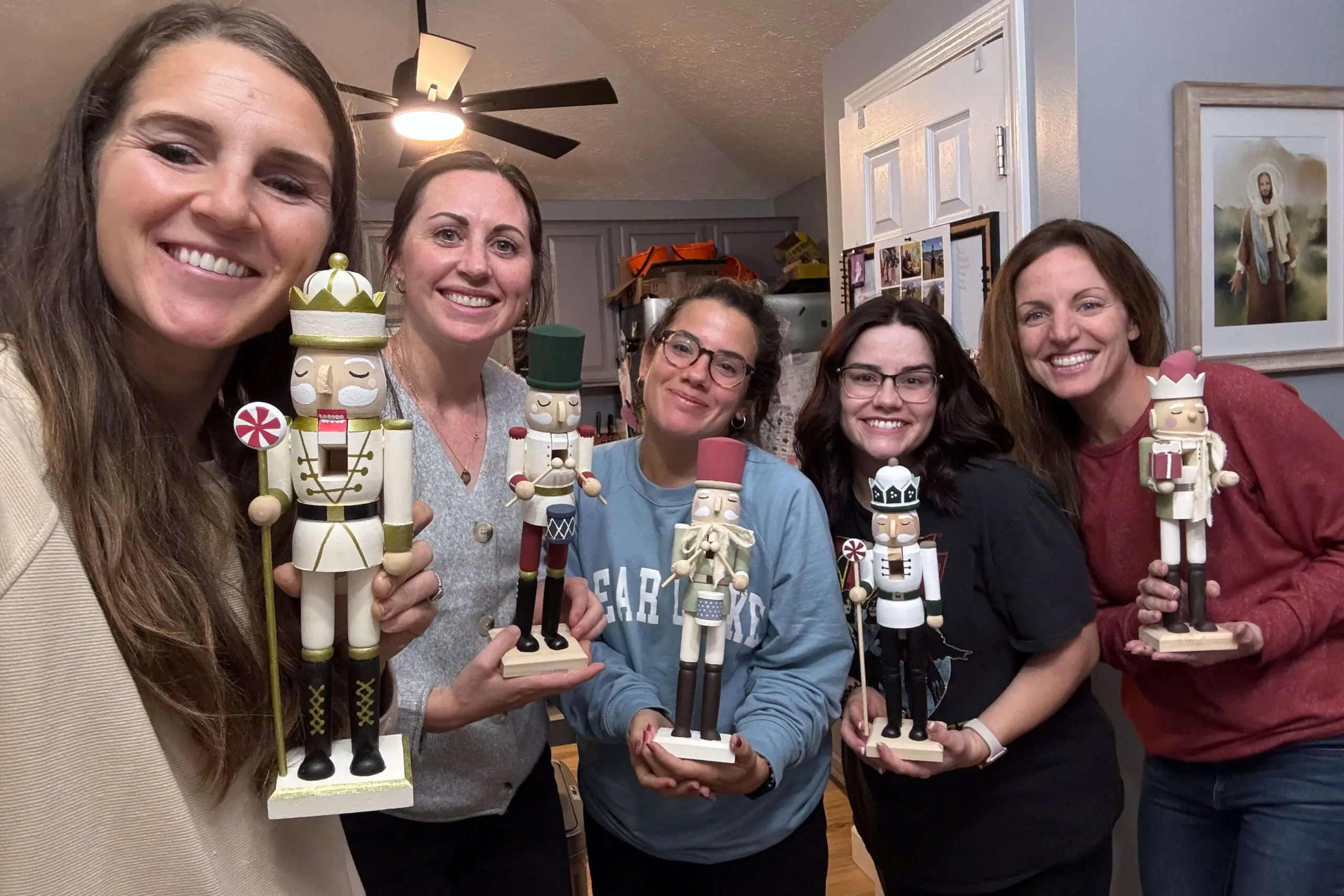 Group of women smiling and holding hand-painted wooden nutcrackers in a cozy kitchen setting during a nutcracker painting session.