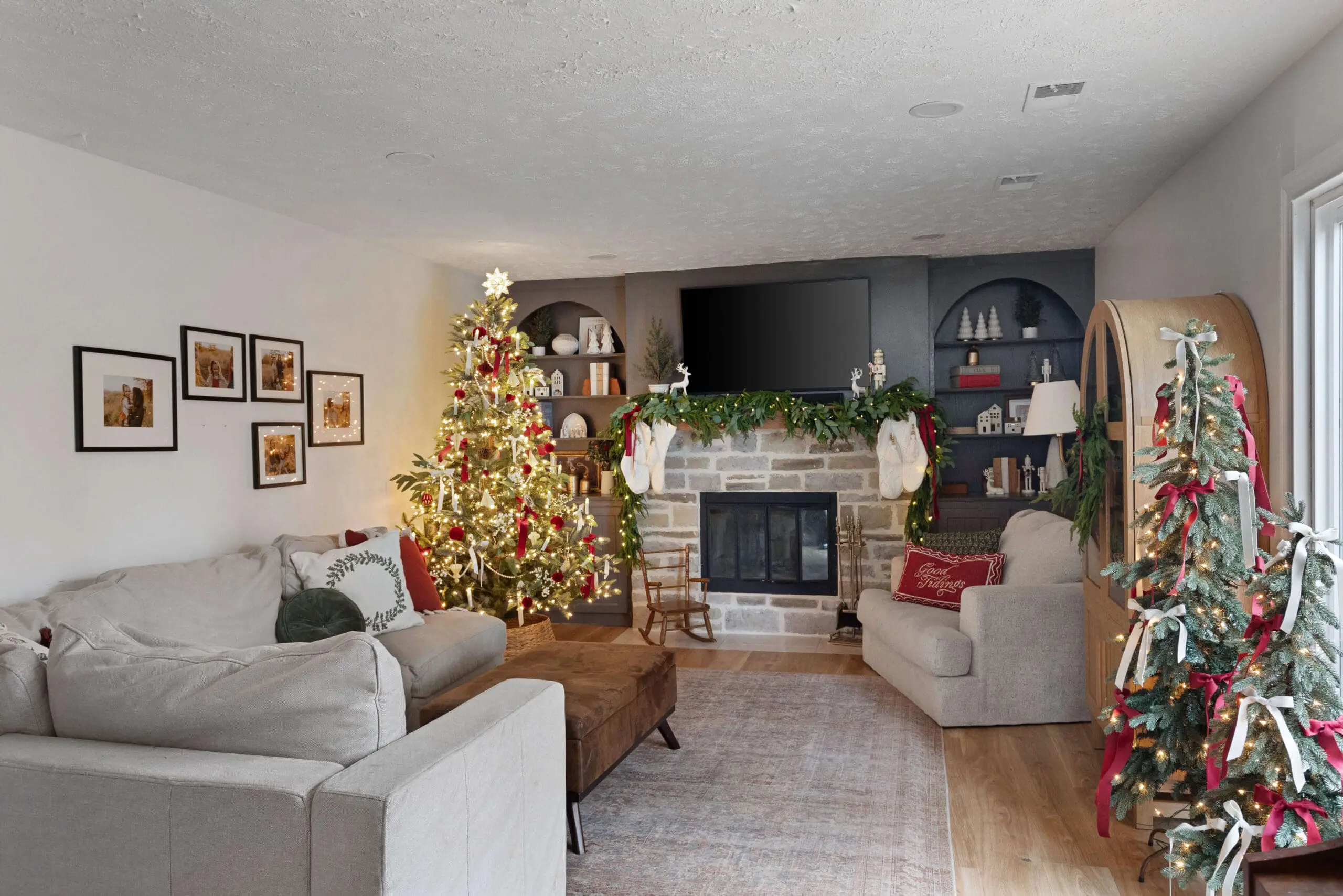 Cozy holiday living room with a large, lit Christmas tree decorated with red and white ornaments, ribbons, and candles. The stone fireplace is adorned with greenery and stockings, while a light gray sectional with festive pillows sits in the foreground, creating a warm, inviting holiday scene.