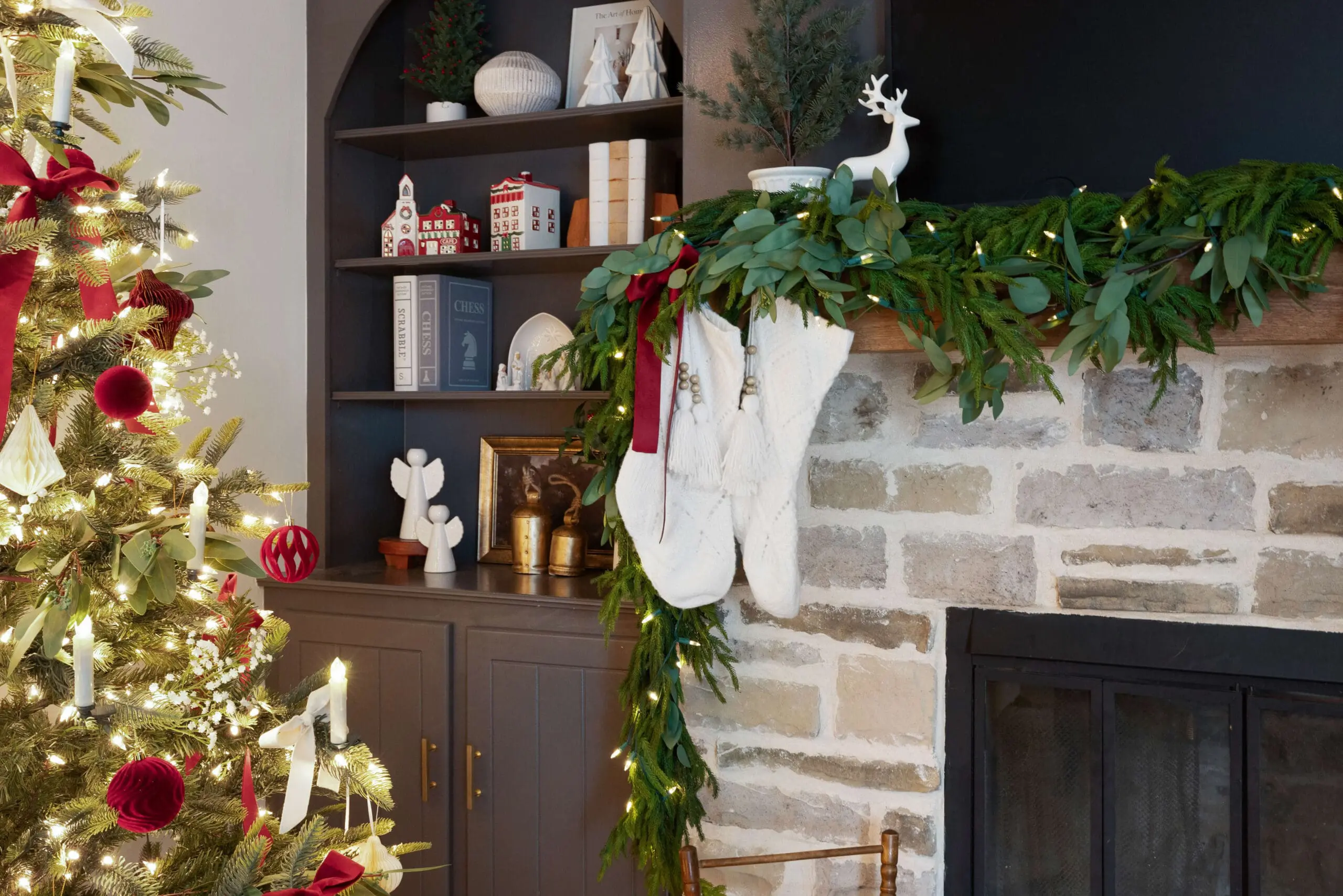 Close-up of a holiday mantel decorated with lush garland, eucalyptus leaves, and a red velvet ribbon. White knit stockings with tassels hang from the mantel, accented by a mini evergreen tree and a ceramic reindeer, enhancing the festive ambiance.