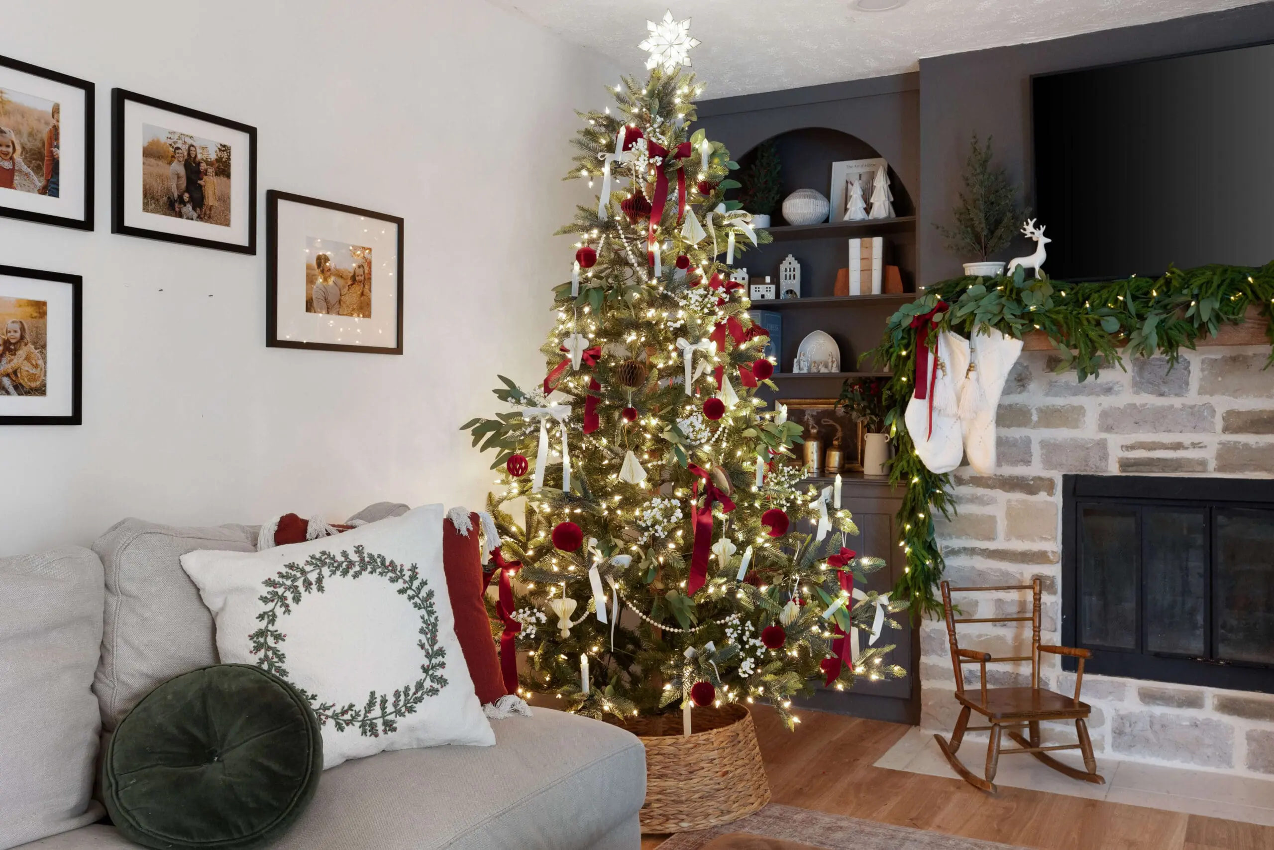 Cozy living room decorated for Christmas with a beautifully adorned tree in red and white tones. The tree, set in a woven basket, features red velvet ornaments, white ribbons, faux greenery, and candle lights. Next to the tree, a stone fireplace is decorated with stockings and a festive garland. Framed family photos hang on the wall above a light gray sofa, accented with holiday-themed pillows, creating a warm and inviting holiday atmosphere.
