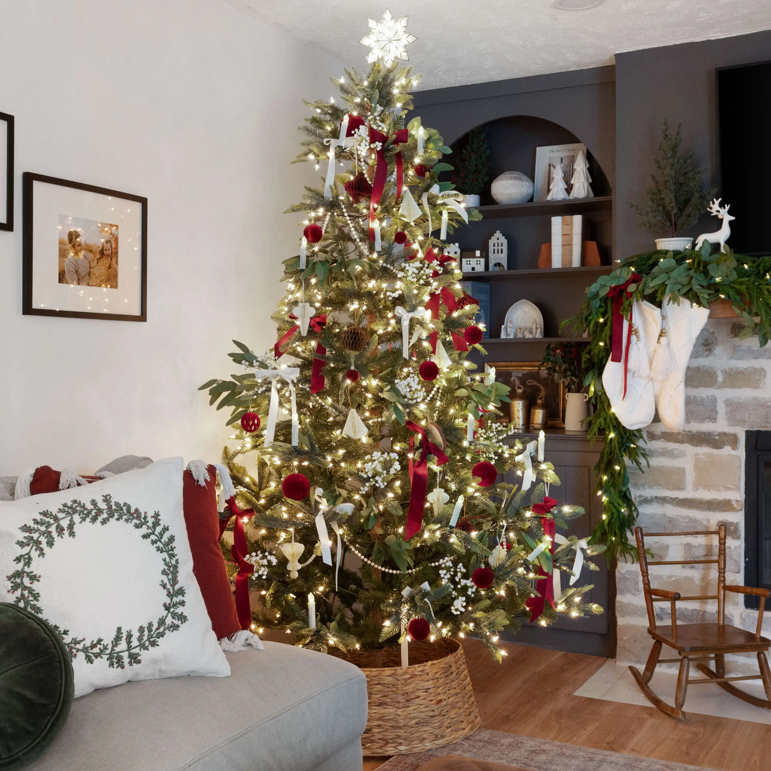 Warm holiday living room featuring an elegant Christmas tree adorned with red velvet ornaments, white ribbons, and candles. The tree is placed in a woven basket and topped with a glowing star. Cozy pillows in red and green accent a light gray couch nearby, while the stone fireplace is decorated with greenery, stockings, and small holiday accents on the shelves. A vintage wooden rocking chair completes the inviting, nostalgic holiday decor.