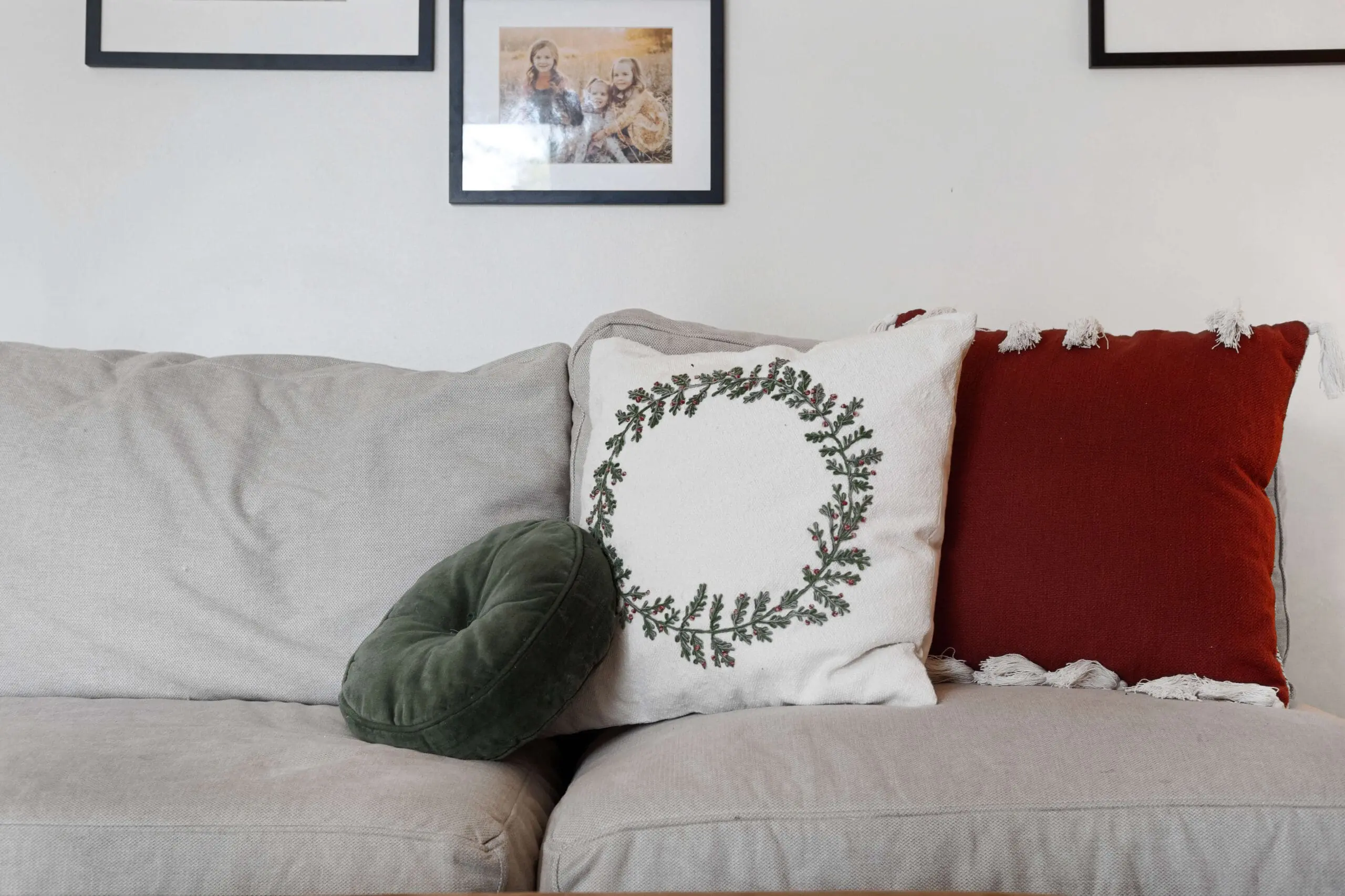 Neutral-toned sofa with holiday-themed accent pillows, including a white pillow featuring a green and red berry wreath design, a deep green velvet round pillow, and a rust-colored pillow with white tassels. Above the sofa, framed family photos add a personal touch to the cozy living room decor.