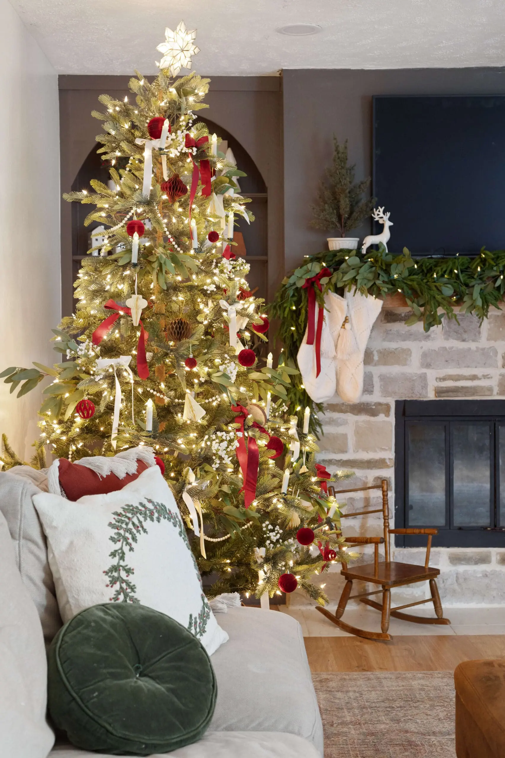 Inviting holiday living room with a tall, beautifully decorated Christmas tree adorned with twinkling white lights, red velvet ornaments, candles, and soft ribbons. The tree is topped with a glowing star, and lush greenery spills over the nearby stone fireplace mantel, which is decorated with white knit stockings and sprigs of eucalyptus. A cozy couch in the foreground is accented with festive pillows in red and green, while a vintage wooden rocking chair sits by the fireplace, completing the warm, nostalgic Christmas home decor.