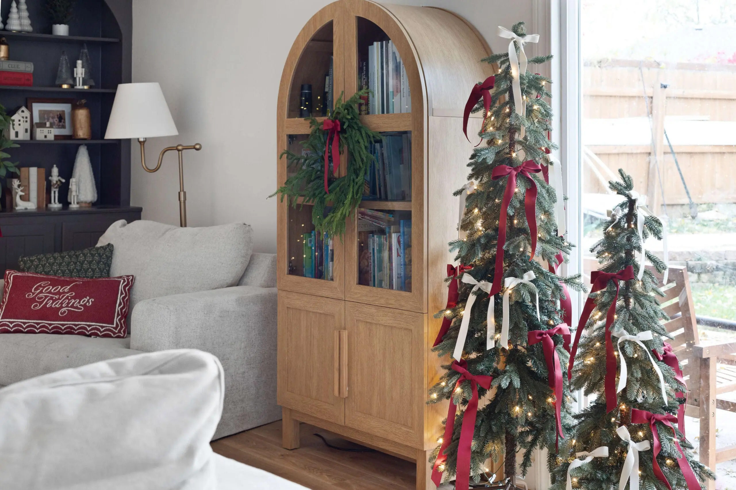 A light wood cabinet with a small wreath on its doors is set beside a pair of slim, lit Christmas trees adorned with red and white ribbons. The cozy corner includes a light gray armchair with festive red and green pillows, adding holiday charm.