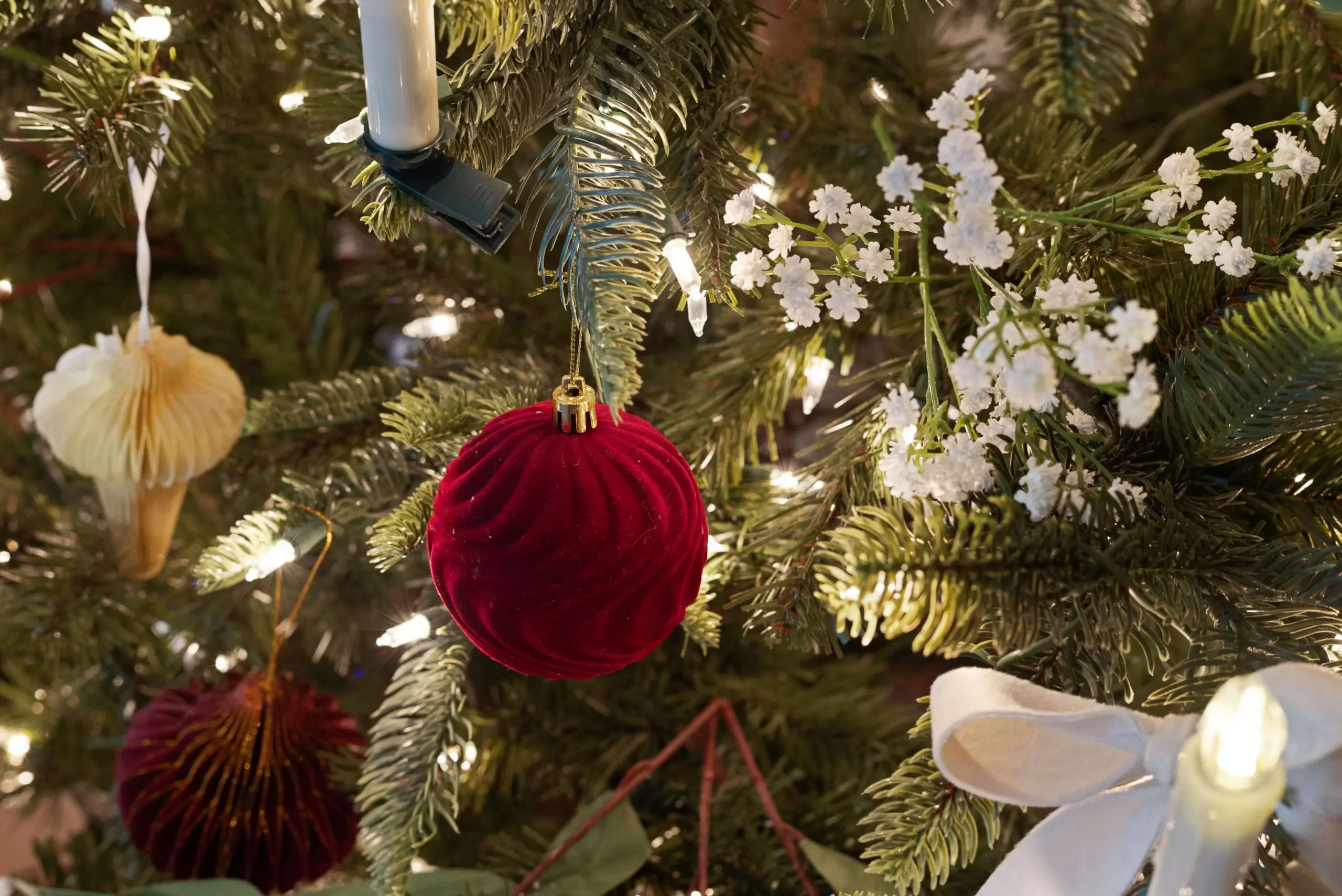 Detailed view of a Christmas tree decorated with a mix of ornaments, including a rich red velvet ball, small white flowers, and hanging candles. The tree’s soft white lights create a magical, warm glow.