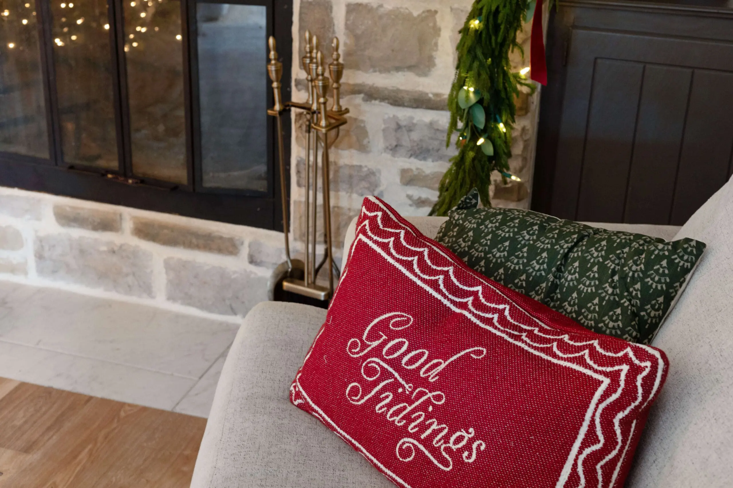 Festive throw pillows on a light gray sofa, featuring a red pillow embroidered with 'Good Tidings' and a green pillow with a subtle holiday pattern. The cozy seating is set beside a stone fireplace, adding a warm touch to the holiday decor.