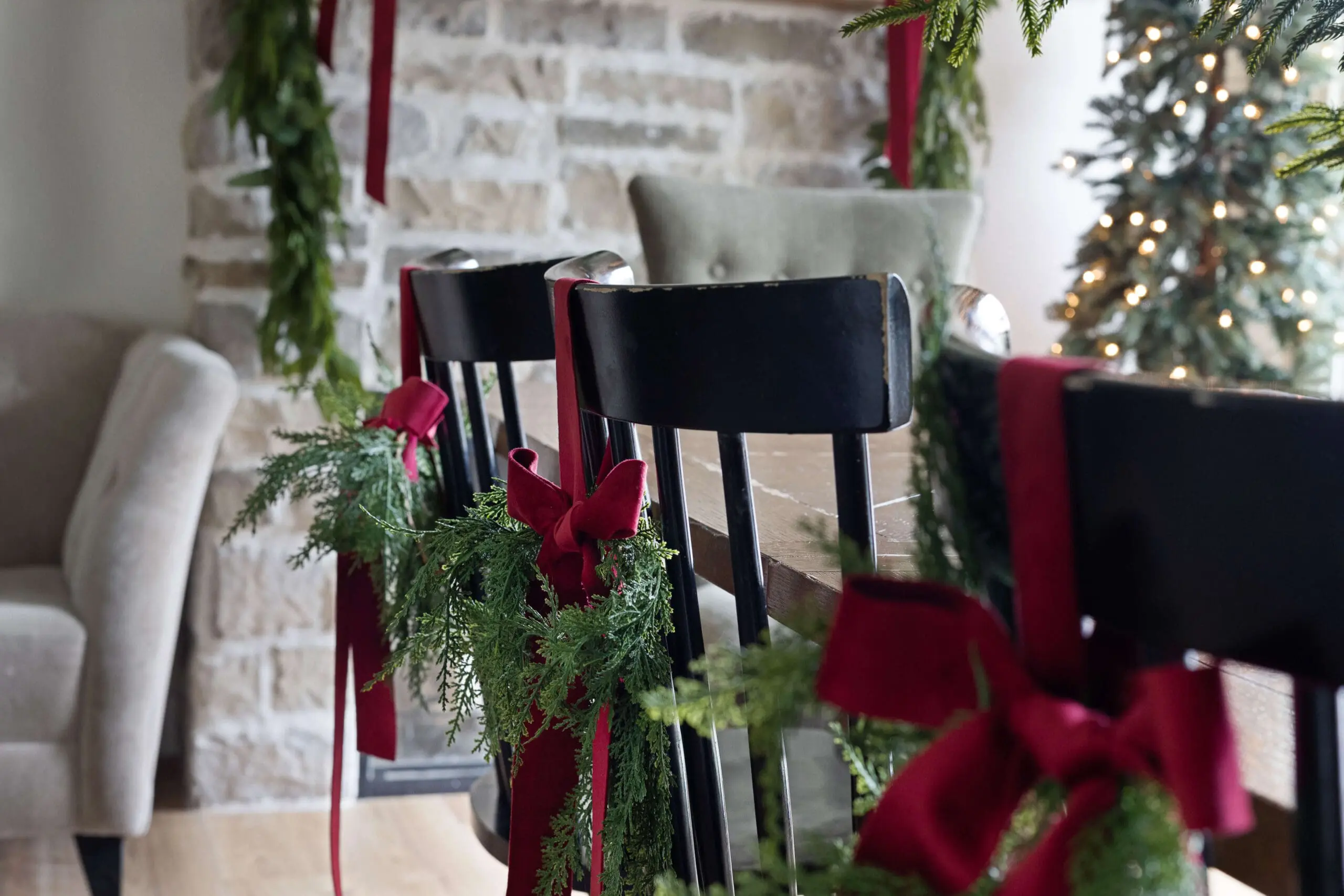 Close-up of black dining chairs dressed up for the holidays with bundles of fresh greenery and rich red velvet ribbons tied to each chair back. The stone fireplace in the background is decorated with a garland, creating cozy Christmas home decor.