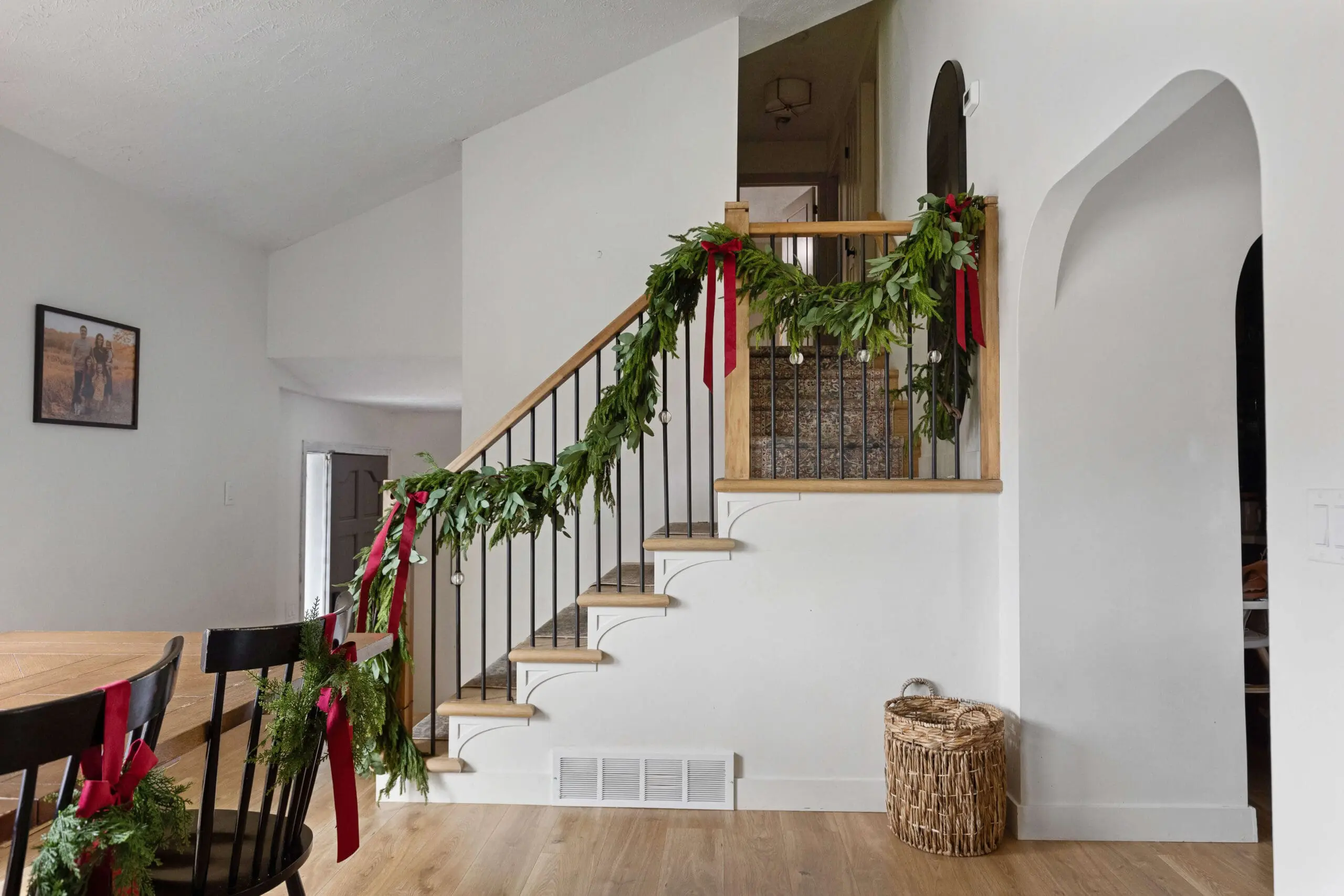Staircase wrapped with a thick greenery garland adorned with red ribbons and silver ornaments. The wood and black metal railing adds a modern rustic touch, while a woven basket near the stairs adds warmth to the neutral decor, completing the Christmas home decor look in this inviting entryway.