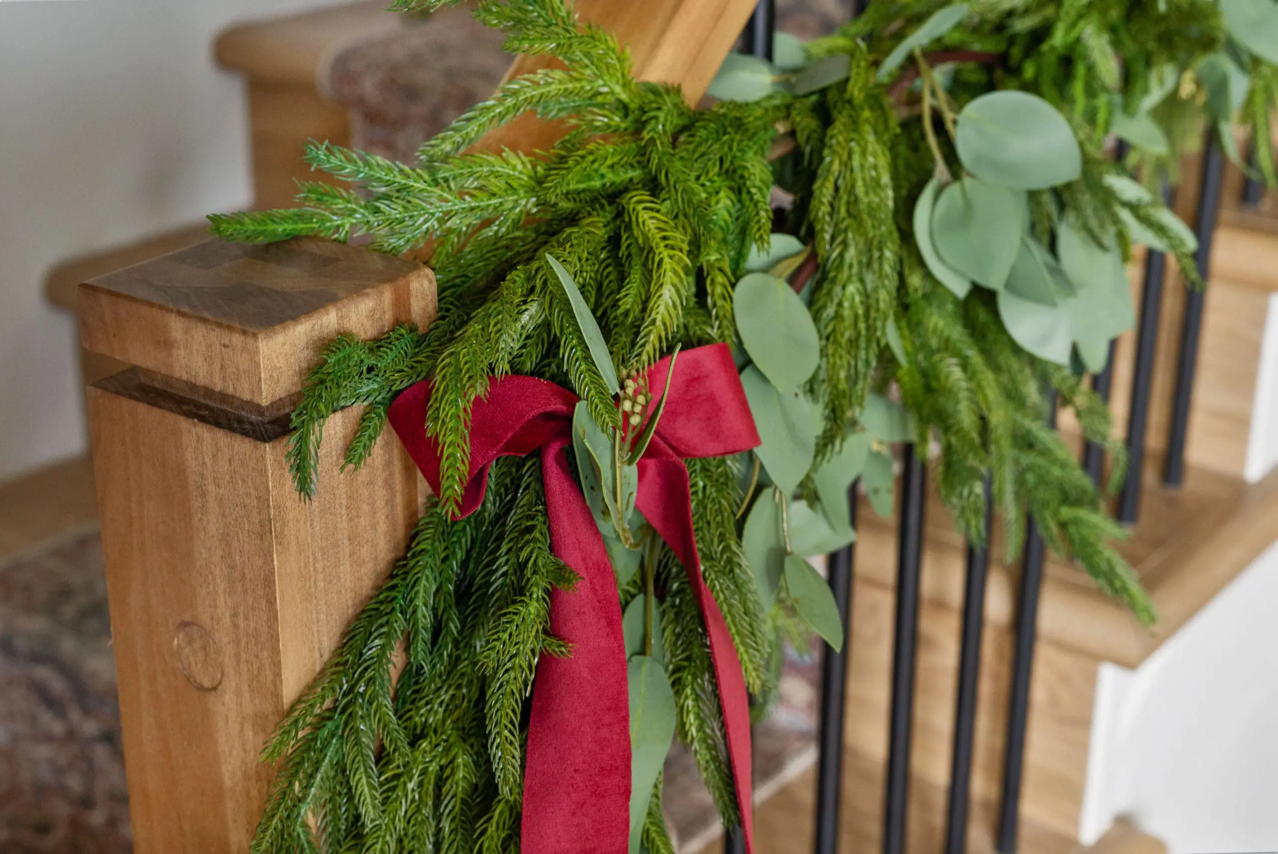 Close-up of a stairway garland made from lush greenery and eucalyptus leaves, accented by a deep red velvet ribbon tied in a bow around the wooden banister post. The natural textures and vibrant colors create a warm, festive feel, adding a touch of elegance to the Christmas home decor.