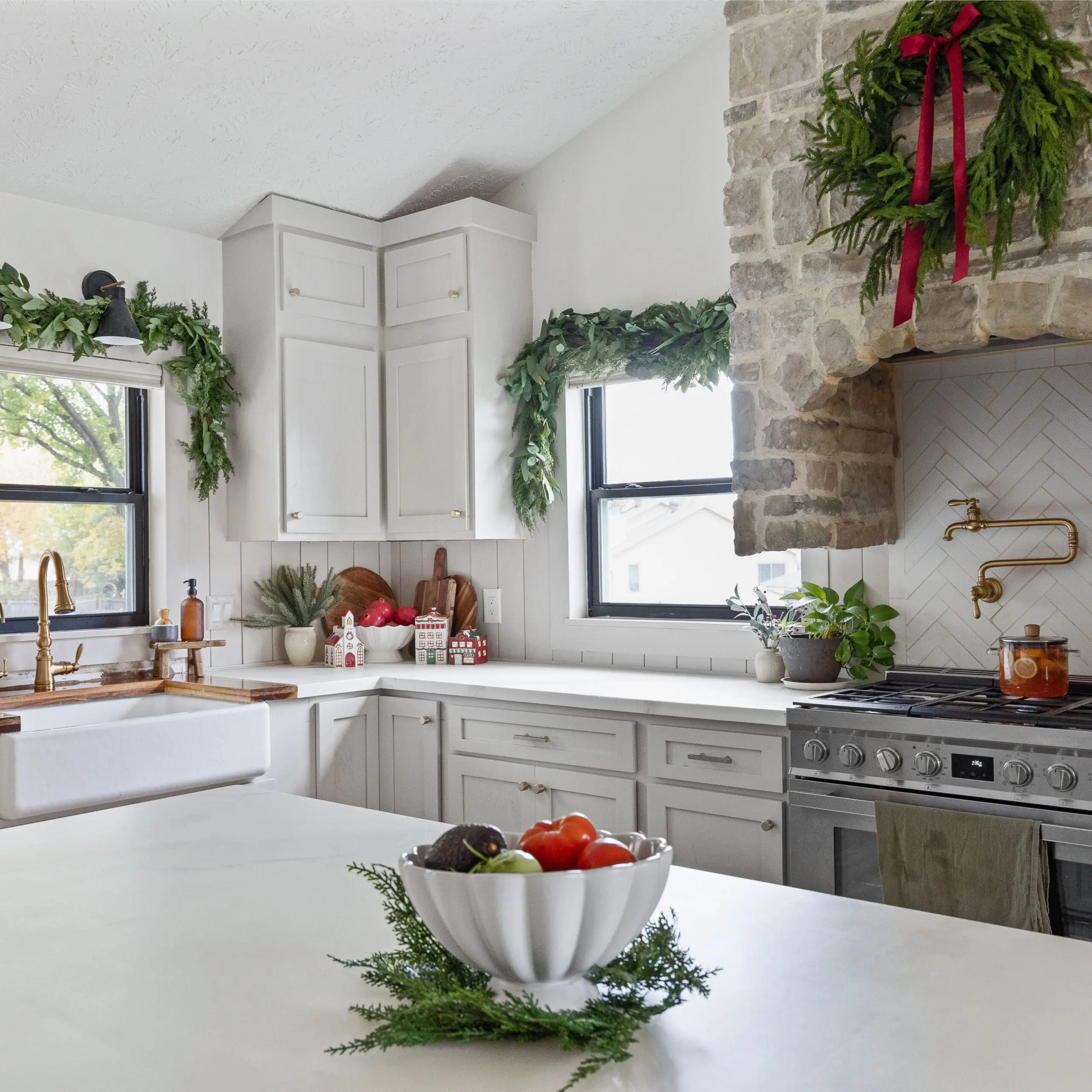 Bright and airy holiday-decorated kitchen with greenery draped over the windows and a large wreath with a red ribbon hanging above a rustic stone range hood. A farmhouse sink with a gold faucet sits below the window, and the countertops feature holiday accents, including a ceramic Christmas village and a bowl of fresh red pomegranates. A white herringbone backsplash and a brass pot filler add a touch of elegance above the stove, where a pot of simmering cider adds warmth to the scene.