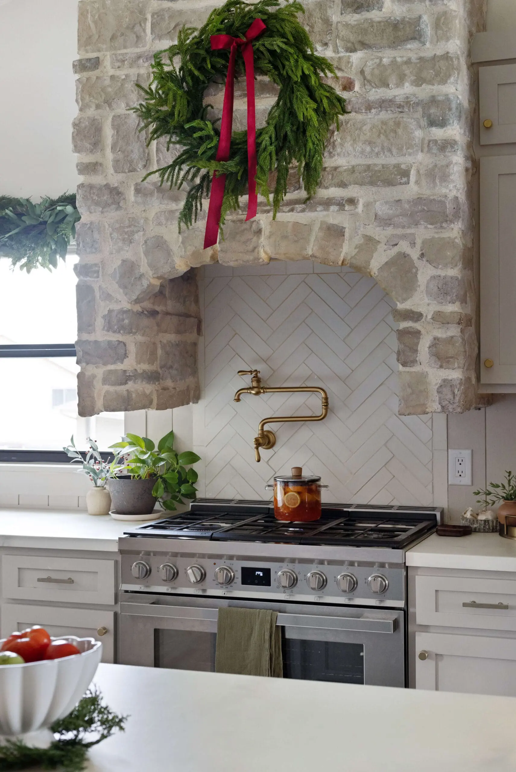 Stone kitchen hood decorated with a lush green wreath tied with a red ribbon, hanging over a stove with a white herringbone-tiled backsplash. The stove features a brass pot filler and a glass pot simmering with citrus slices, adding a cozy touch. Potted plants sit on the white countertop near the window, enhancing the natural and festive feel of the space.