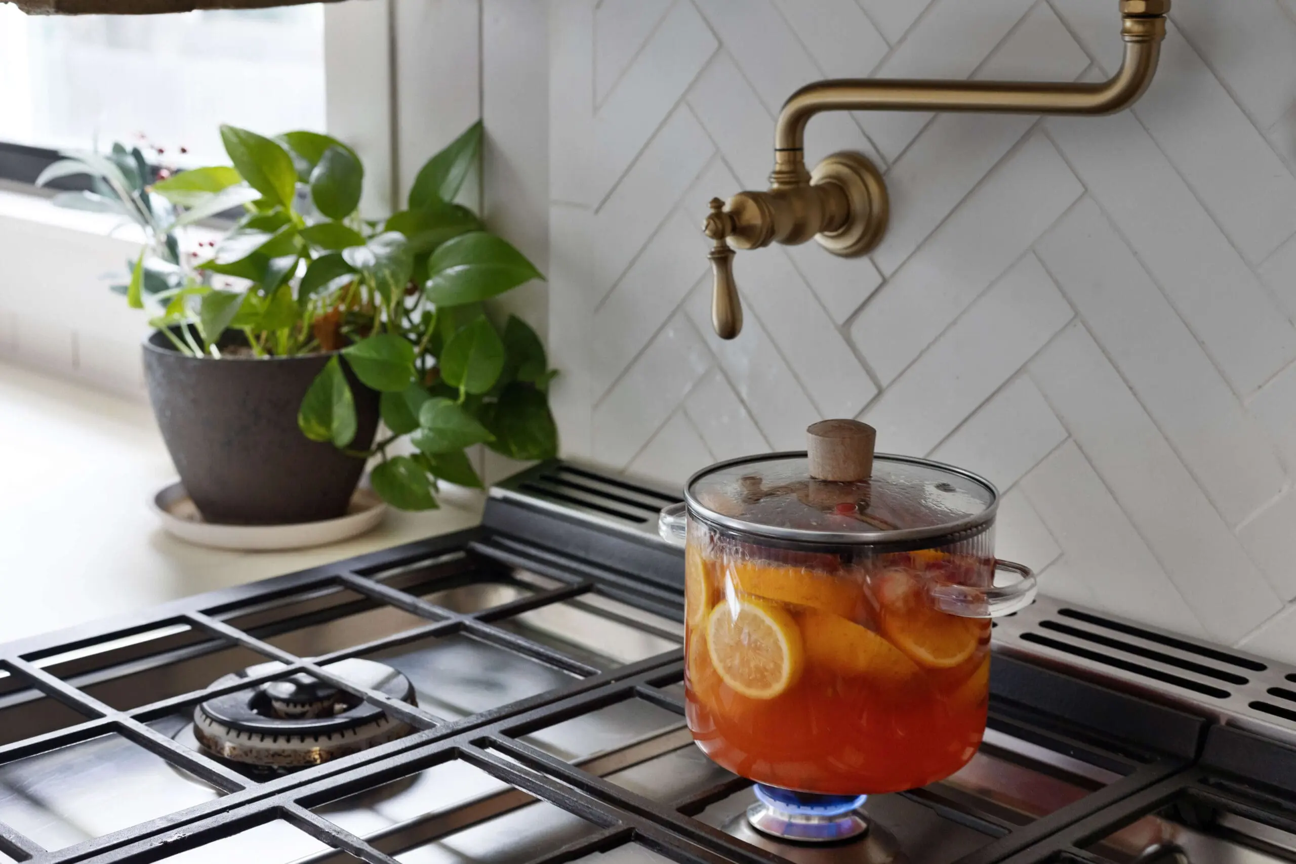 Close-up of a stovetop with a pot of simmering spiced cider, featuring slices of orange and cinnamon sticks, creating a warm and festive aroma. The glass pot is placed on a gas burner with a gentle flame, and above it, a brass pot filler is mounted on a white herringbone tile backsplash. A potted green plant adds a touch of freshness beside the stove, enhancing the inviting, seasonal kitchen atmosphere.