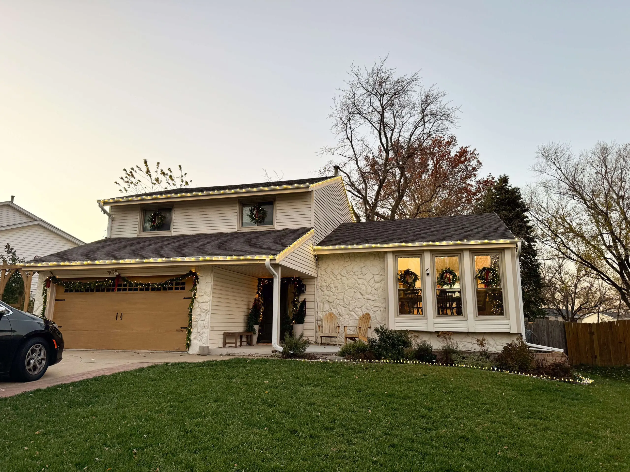 A beautifully decorated house at dusk with warm white string lights outlining the roofline and garage, wreaths in the windows, and festive greenery garland framing the entrance. A lush green lawn completes the cozy holiday scene.