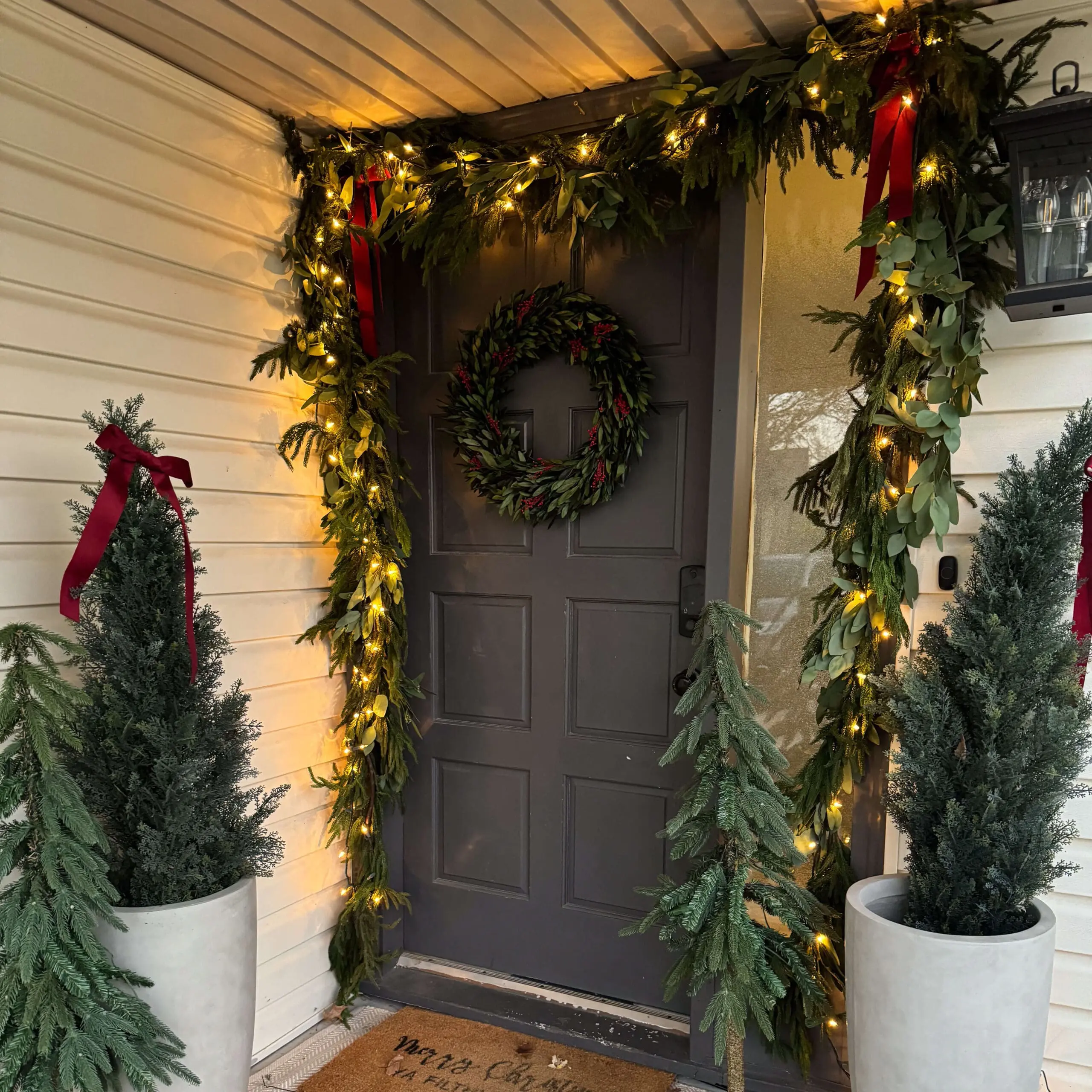 A close-up of a front door adorned with twinkling string lights and fresh greenery garland, featuring festive red bows and a holiday wreath with red accents. Tall potted evergreens and small artificial Christmas trees flank the door, creating a warm and inviting entryway.