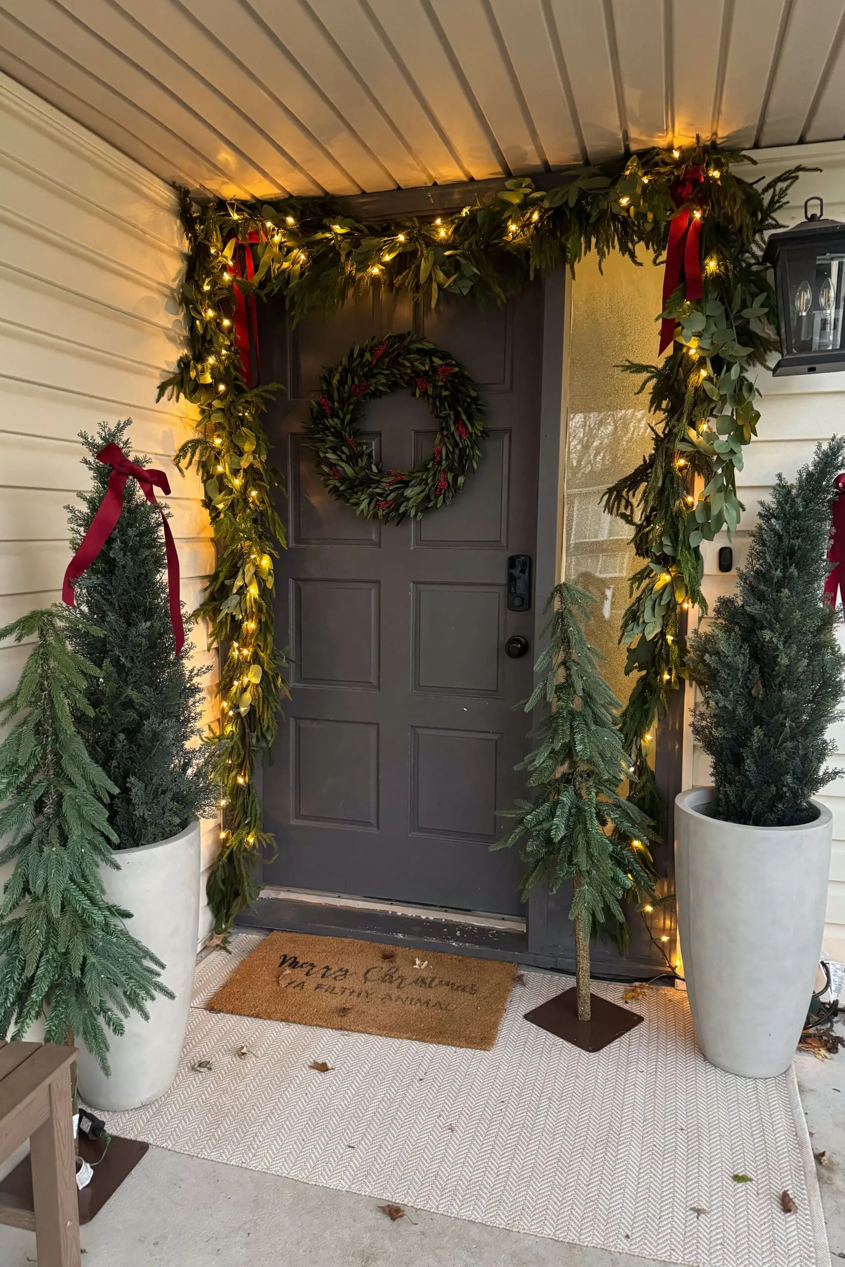 A front porch fully decorated for the holidays with glowing string lights, greenery garland, red bows, and a wreath on a gray door. The setup includes potted evergreens and small artificial Christmas trees, complemented by a humorous holiday-themed doormat.
