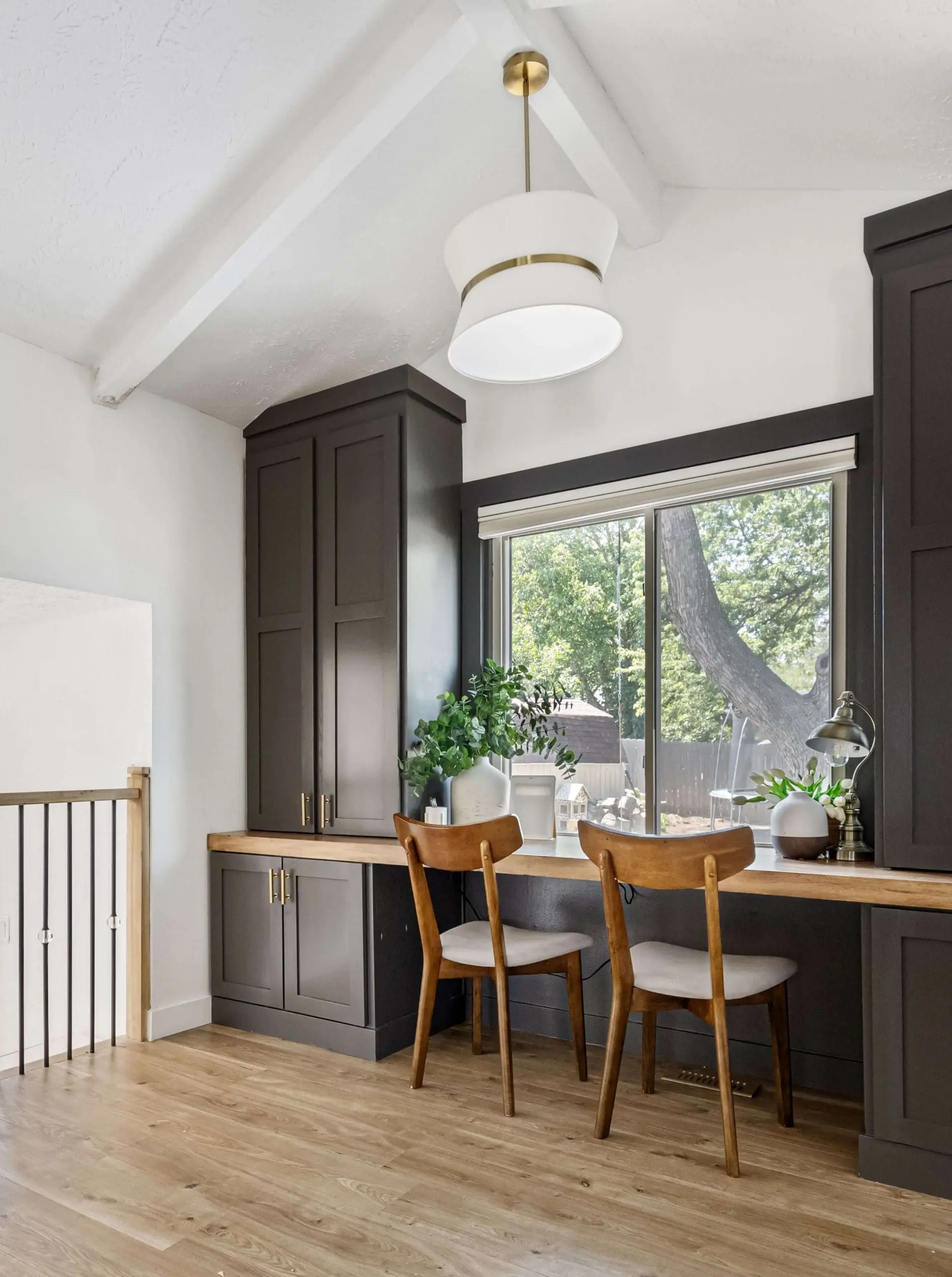 Bright and inviting home workspace with dark cabinetry, a wooden countertop, and two mid-century modern chairs. A large window offers a view of a lush backyard, while simple greenery and minimal decor create a clean, functional aesthetic under a modern pendant light.