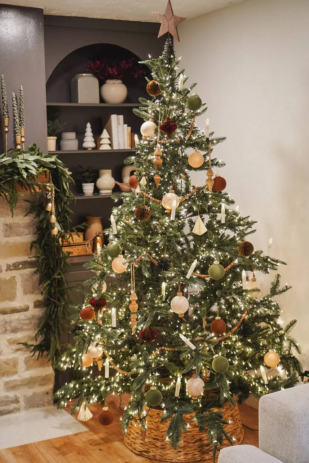 A beautifully decorated Christmas tree in a cozy living room featuring warm neutral and earthy-toned ornaments, including wooden shapes, velvet balls, and gold accents. The tree is illuminated with soft white lights and topped with a wooden star. A nearby stone fireplace is adorned with greenery, and built-in shelves display festive ceramic trees and neutral holiday décor.