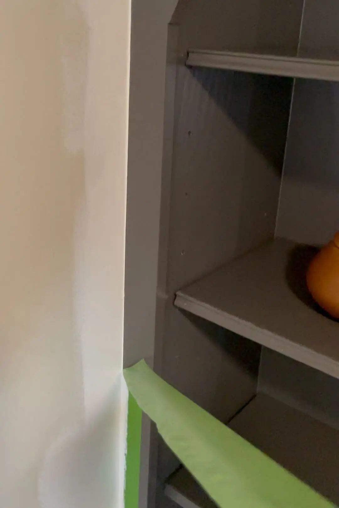 Close-up of a freshly painted beige wall with green painter’s tape neatly applied along the edge, separating the wall from a built-in shelf painted in a dark gray color, partially visible with a pumpkin as decor.