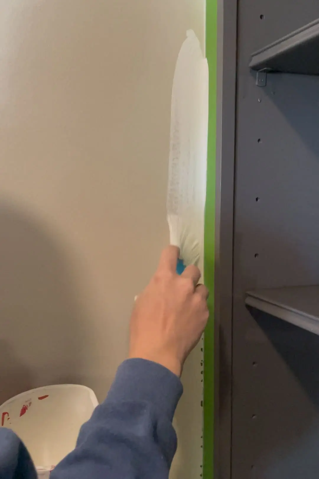 A hand using a brush to apply white paint along the same taped-off edge of a built-in shelving unit, ensuring a smooth and professional finish during the DIY project.

