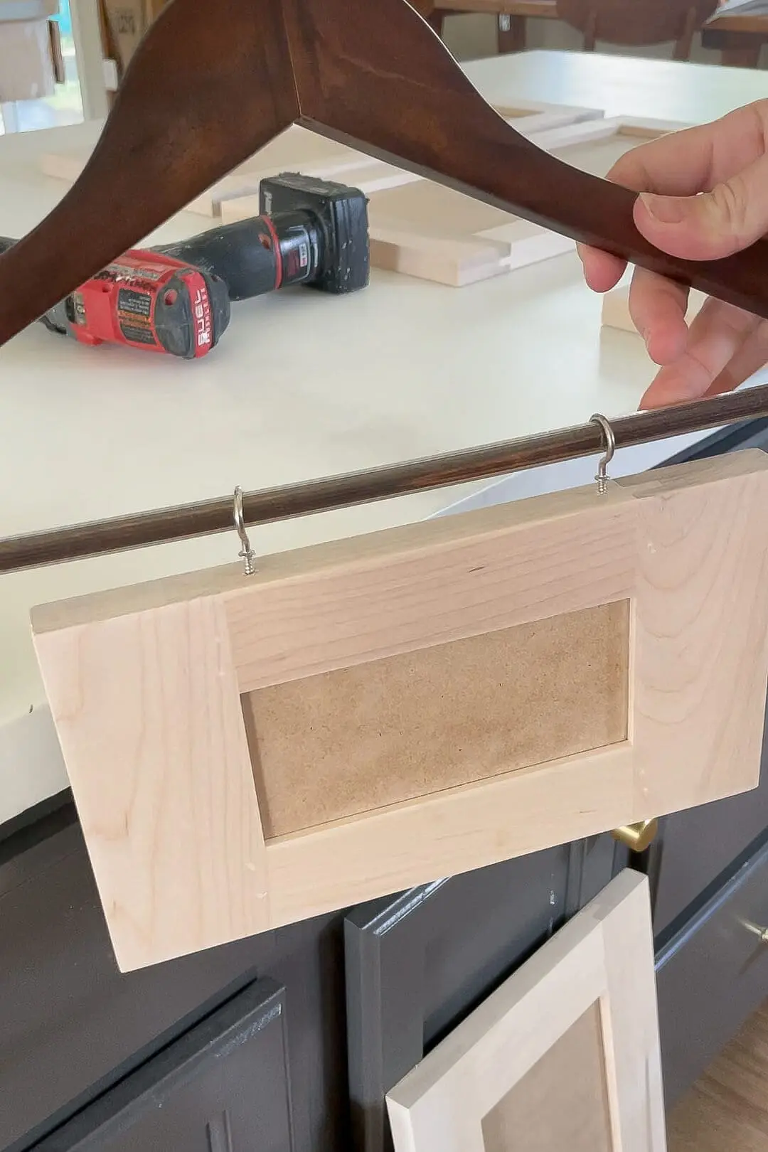Close-up of a wooden cabinet door frame attached to a dark wooden hanger using metal hooks, with a power drill and other wooden pieces on a work surface in the background.
