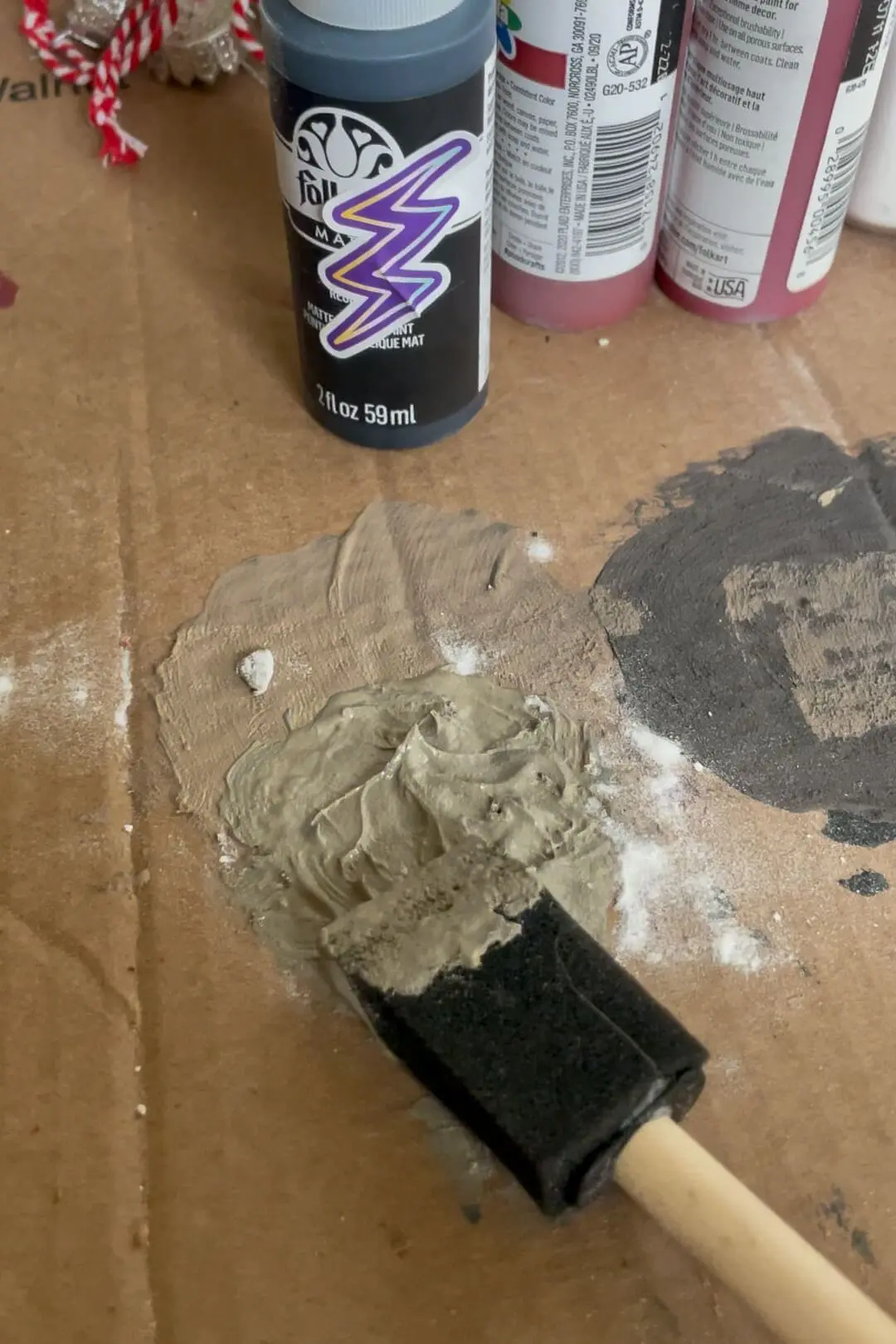 A bottle of FolkArt matte acrylic paint with gray paint and a foam brush on a cardboard surface, used for crafting.