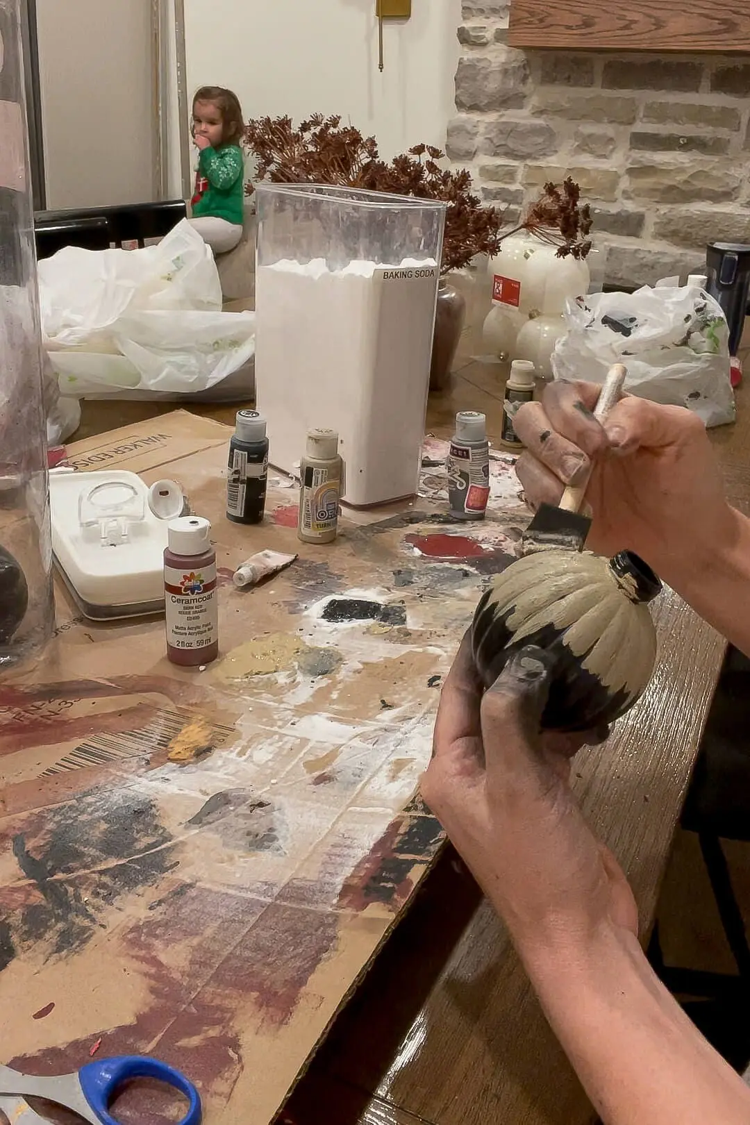 Hands painting a black ornament with beige paint using a foam brush, with various craft supplies, paint bottles, and baking soda visible on a table in the background. 