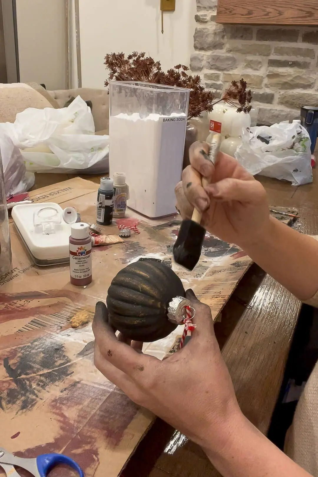 A person painting a black ornament with a foam brush at a crafting table, surrounded by various paint bottles, baking soda, and crafting materials. The ornament is tied with a red and white striped string. This is a faux flocked Christmas ornament.