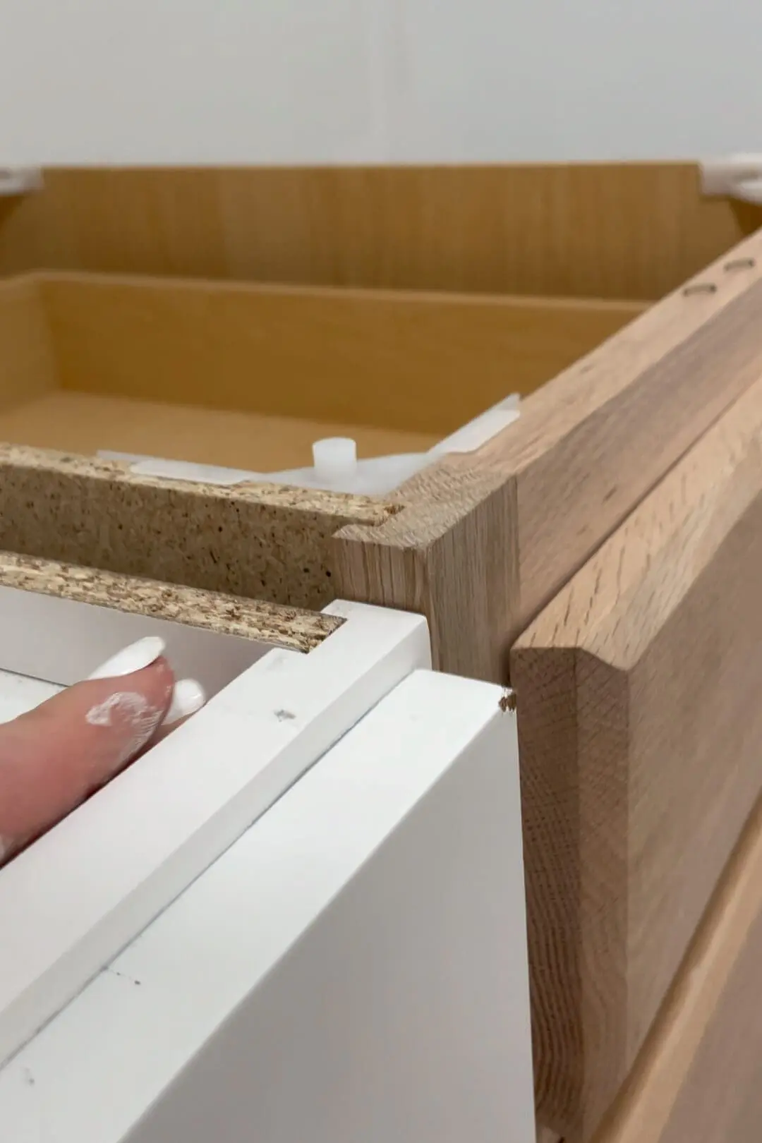 Detailed shot of the seam where a white bathroom cabinet meets an unfinished wood drawer cabinet. A hand with white paint smudges is holding the cabinet edge, highlighting the construction material and alignment during installation.