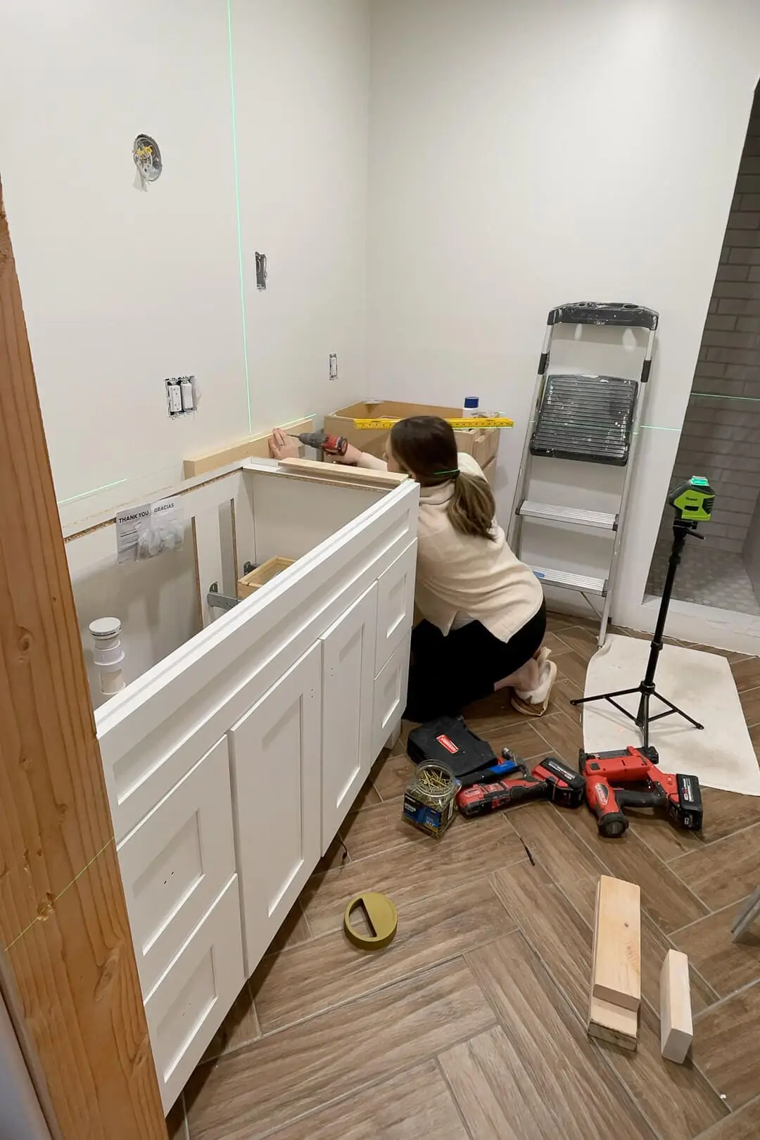 A woman installing a white shaker-style bathroom cabinet during renovation. Tools, a ladder, and a green laser level are spread across a wood-look herringbone floor. The unfinished wood cabinet is partially installed in the background.