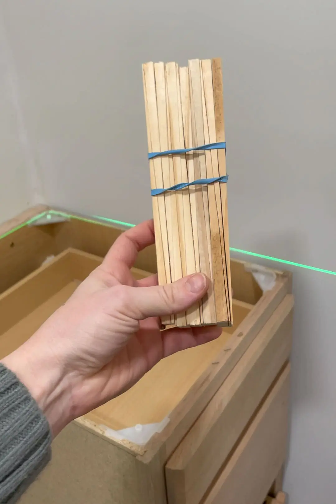 Close-up of a hand holding a bundle of wooden shims tied with blue rubber bands, with an unfinished wooden cabinet and a green laser level line visible in the background.